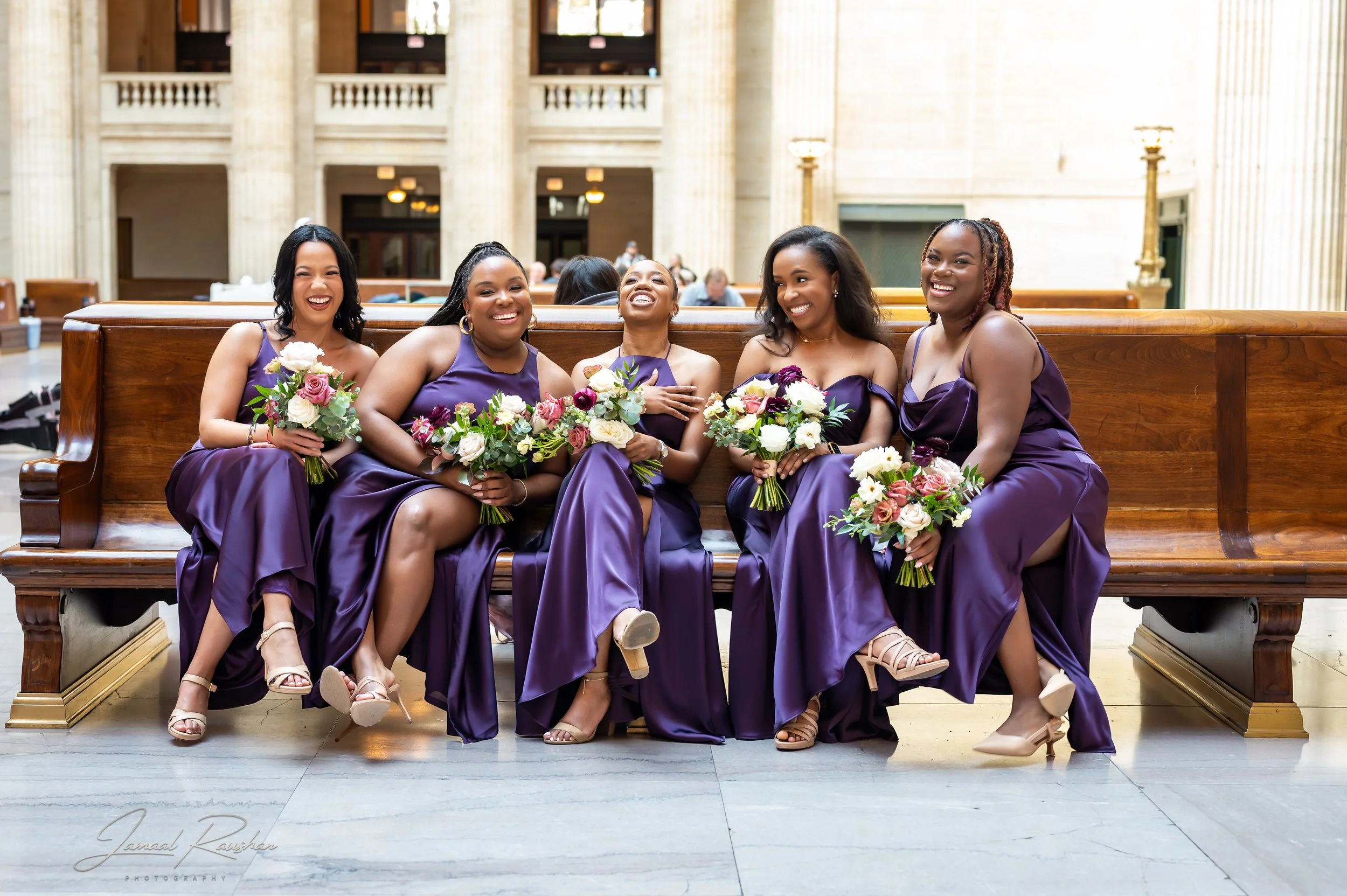 A group of five women wearing matching purple dresses, sitting on a wooden bench inside a courthouse or church, holding bouquets of flowers, and smiling.