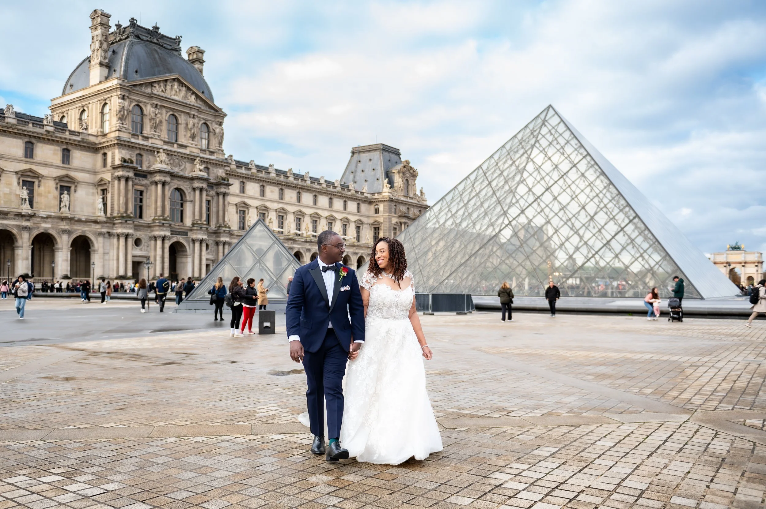 A newlywed couple walking and holding hands in front of the Louvre Museum in Paris, France. The groom is dressed in a navy tuxedo with glasses, and the bride is wearing a white lace wedding gown with curly hair.