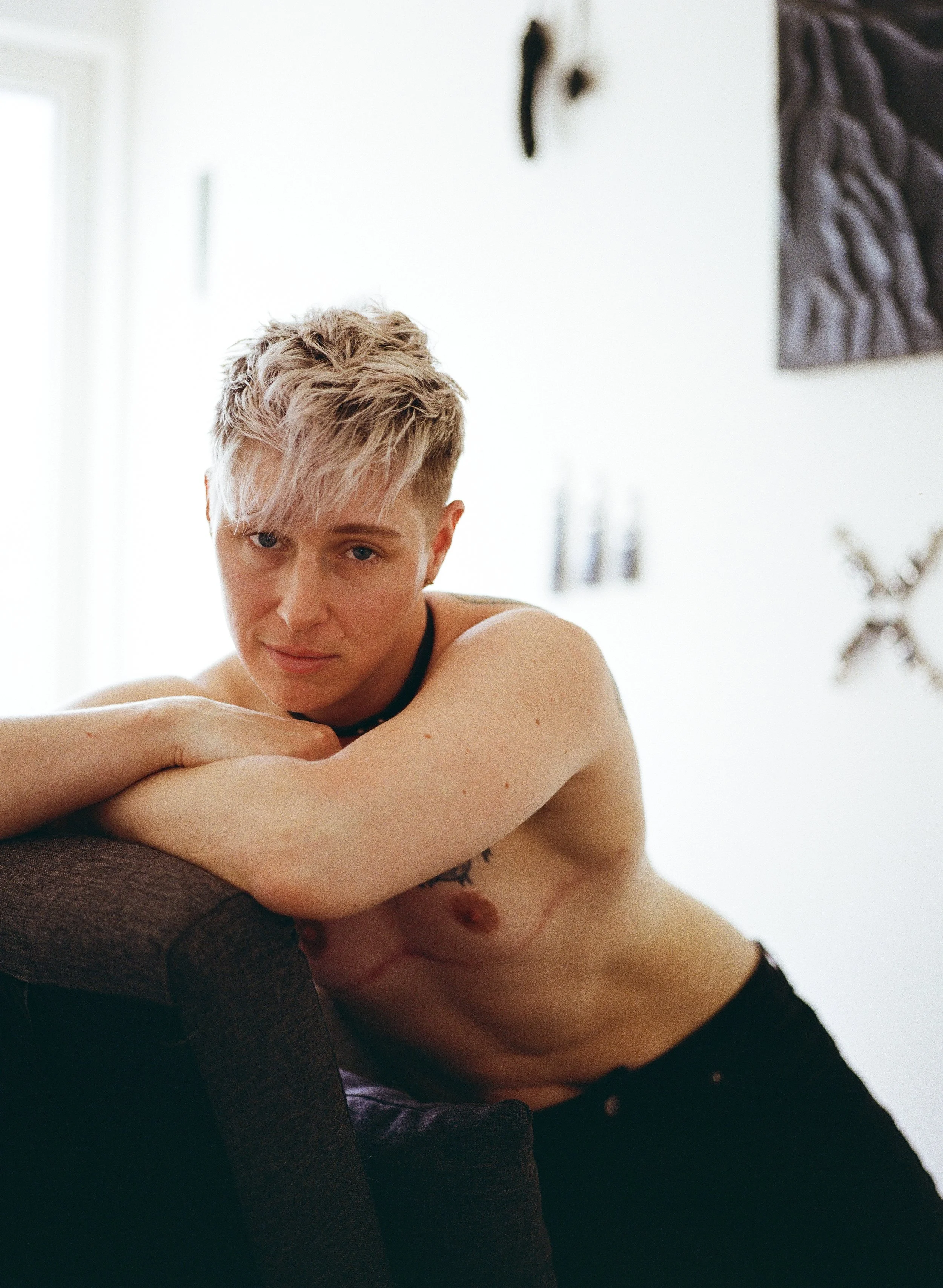 A photo of Jaq Quicksilver, shirtless in black jeans, leaning over a couch and smiling at the camera.