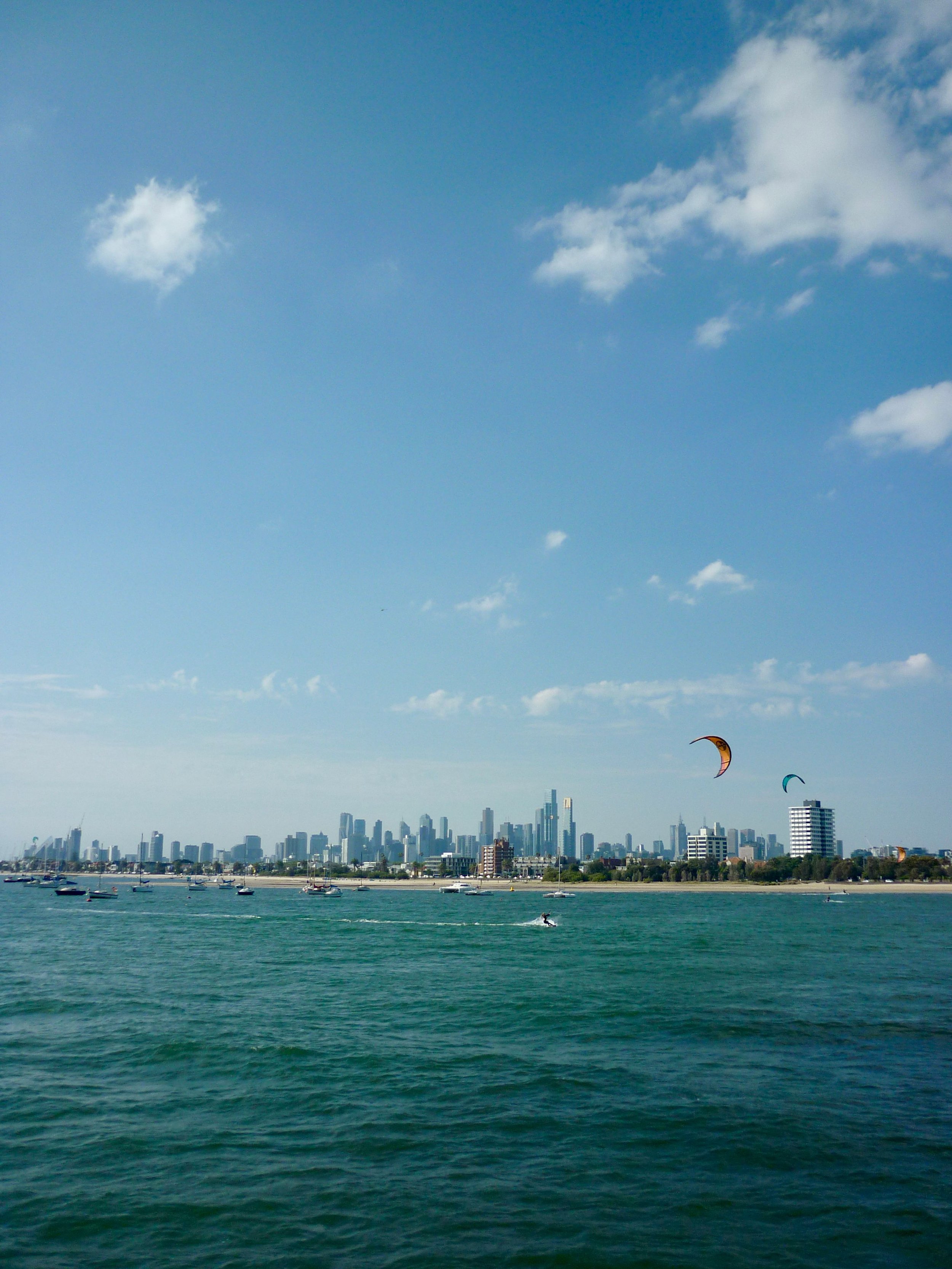 A city skyline with tall buildings, boats on the water, and kite surfers with colorful kites in the sky, under a partly cloudy blue sky.