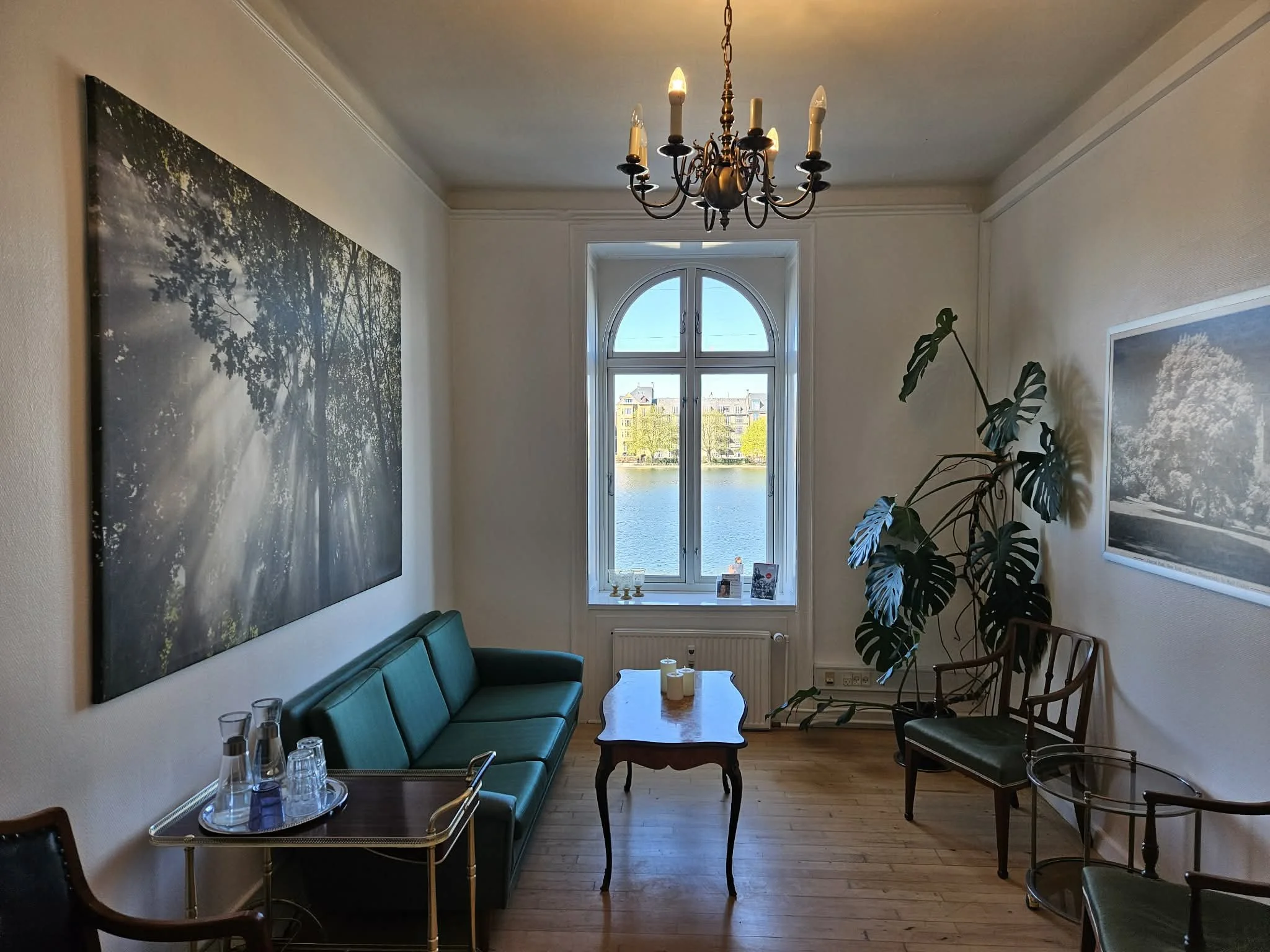 Et hyggeligt venteværelse med grøn sofa, et rundt bord, store vægkunstværker, store vinduer med udsigt til søer og bybaggrund, en lysekrone i loftet, og et stort bladet plante.