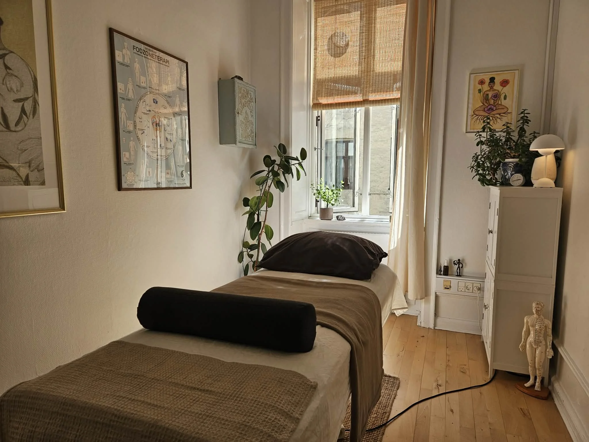 Indendørs billede af et kliniklokale med en massagebriks, grønne planter ved vinduet, vægdekorationer og en kommode med pynt og en lampe