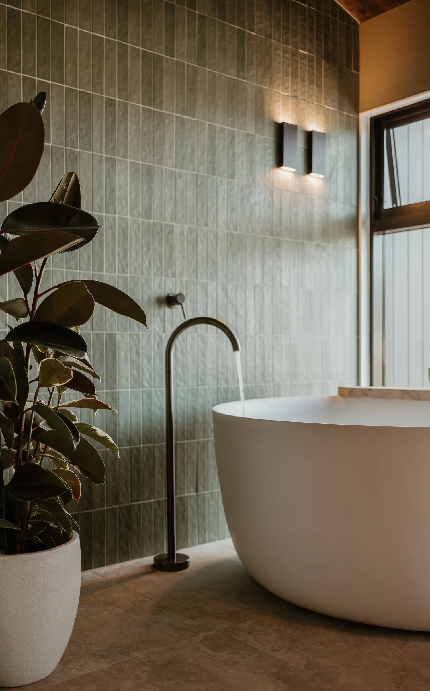 Modern bathroom features a white freestanding bathtub with a wooden tray, a green tiled wall, black floor-standing faucet, a potted plant with dark green leaves, a window, and wall-mounted light fixtures.