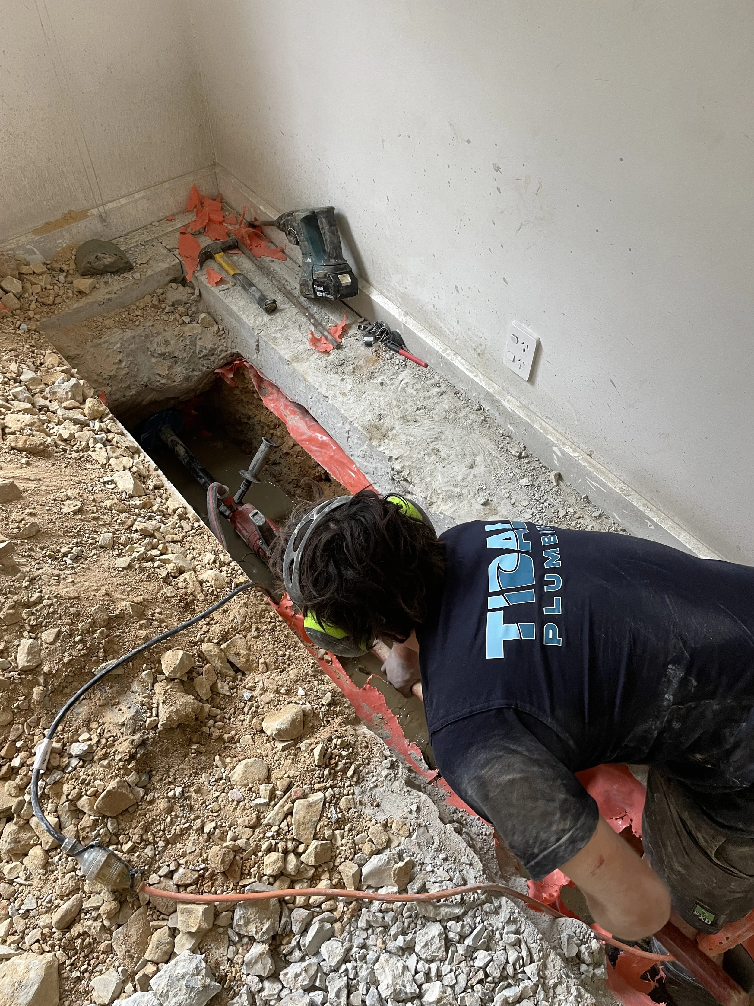 A worker wearing a helmet and a shirt with 'Trinity Plumbing' working on installing or repairing pipes in a trench in a room with dirt and construction tools