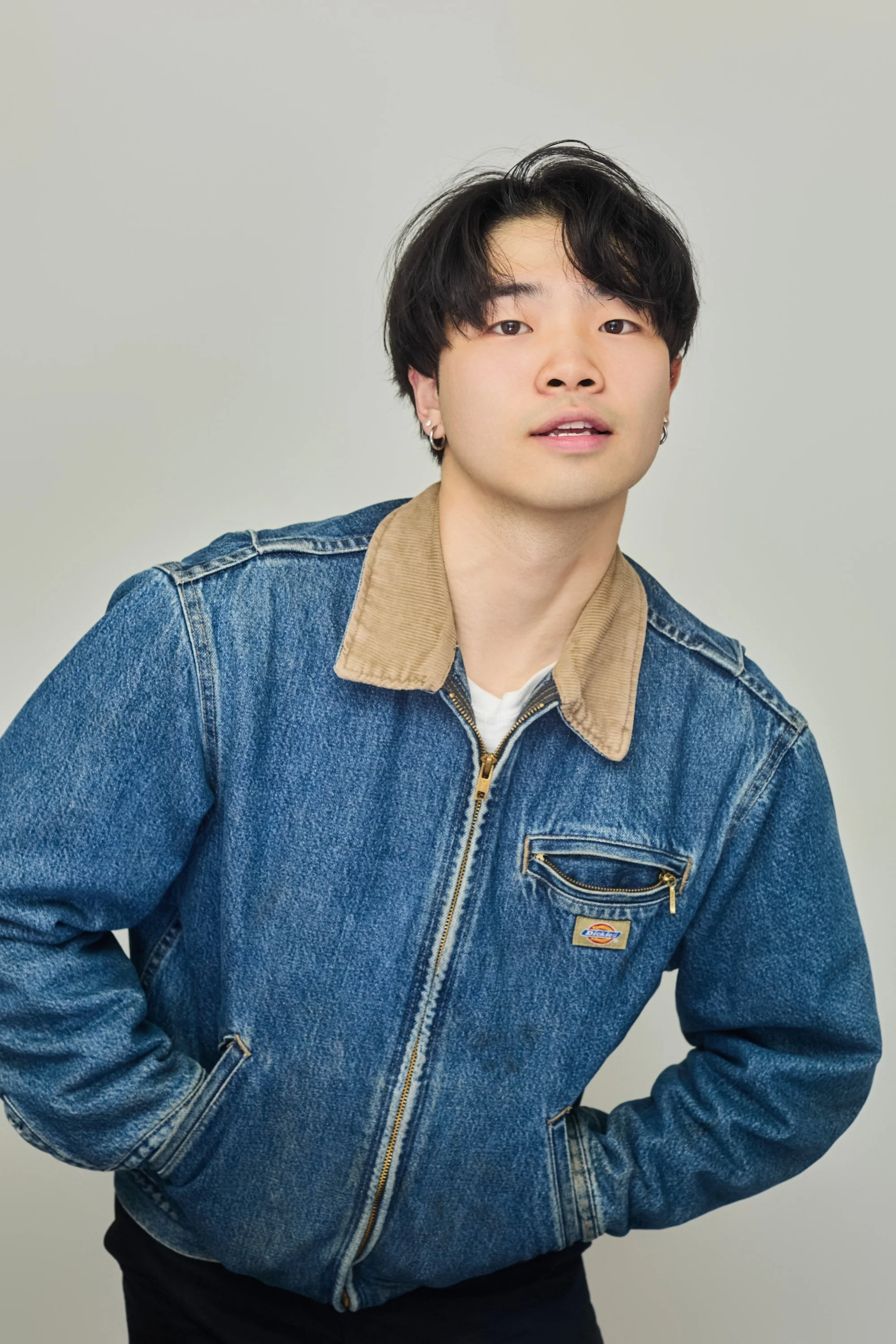 A young man with black hair and earrings, wearing a denim jacket with a tan collar, stands against a plain background.