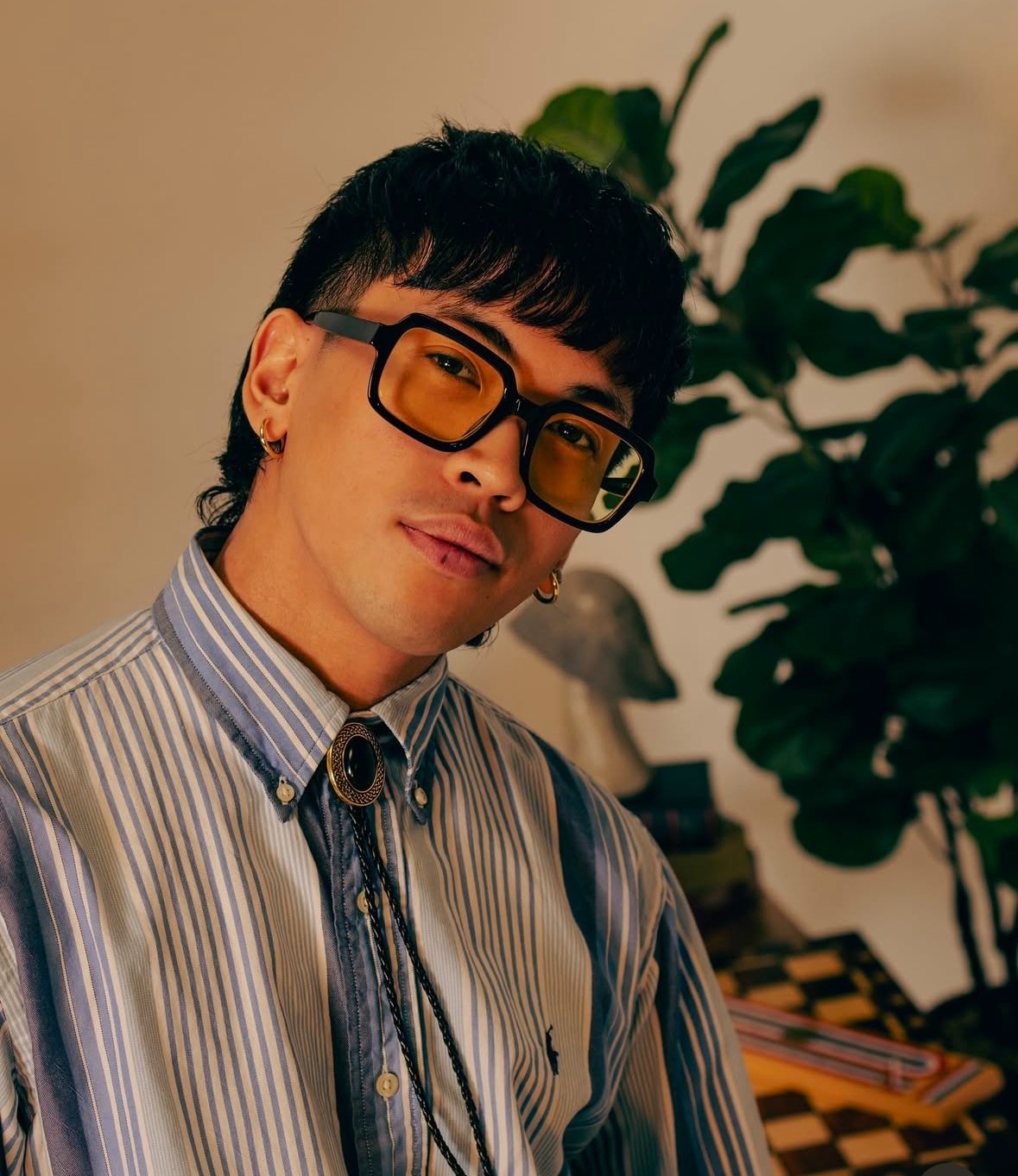 A person with short black hair, wearing oversized square yellow-tinted glasses, earrings, and a striped button-up shirt with a bolo tie, posing indoors with a green leafy plant in the background.