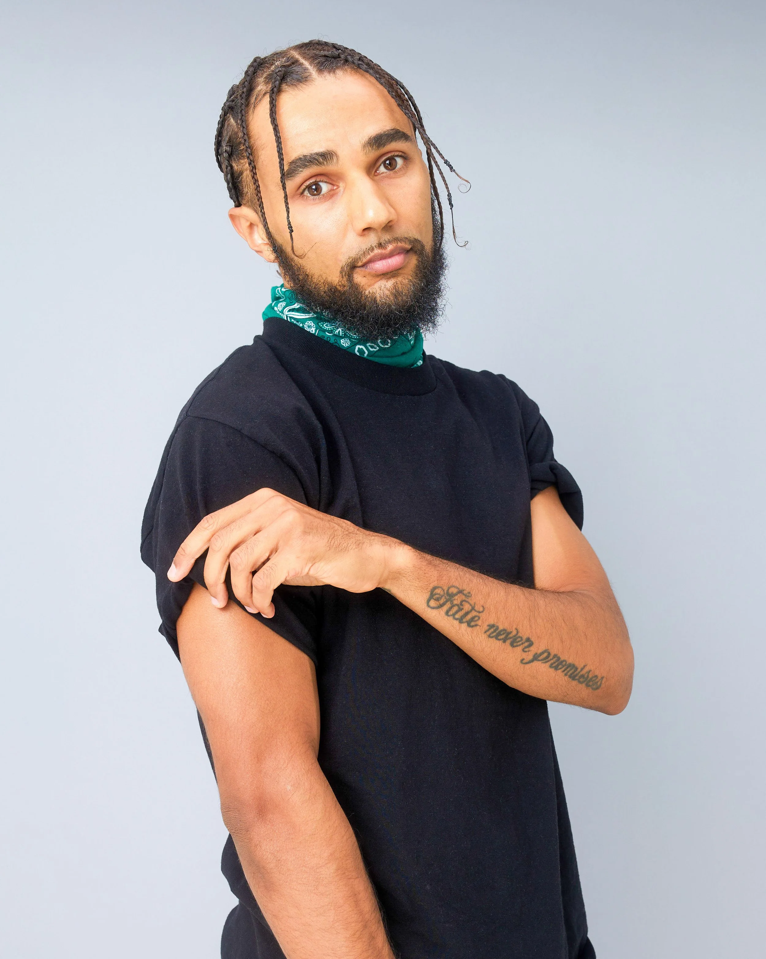 A young man with braided hair and a beard, wearing a black t-shirt and a green bandana around his neck, posing with one arm bent to show a tattoo that reads 'Fate never promises' on his forearm.