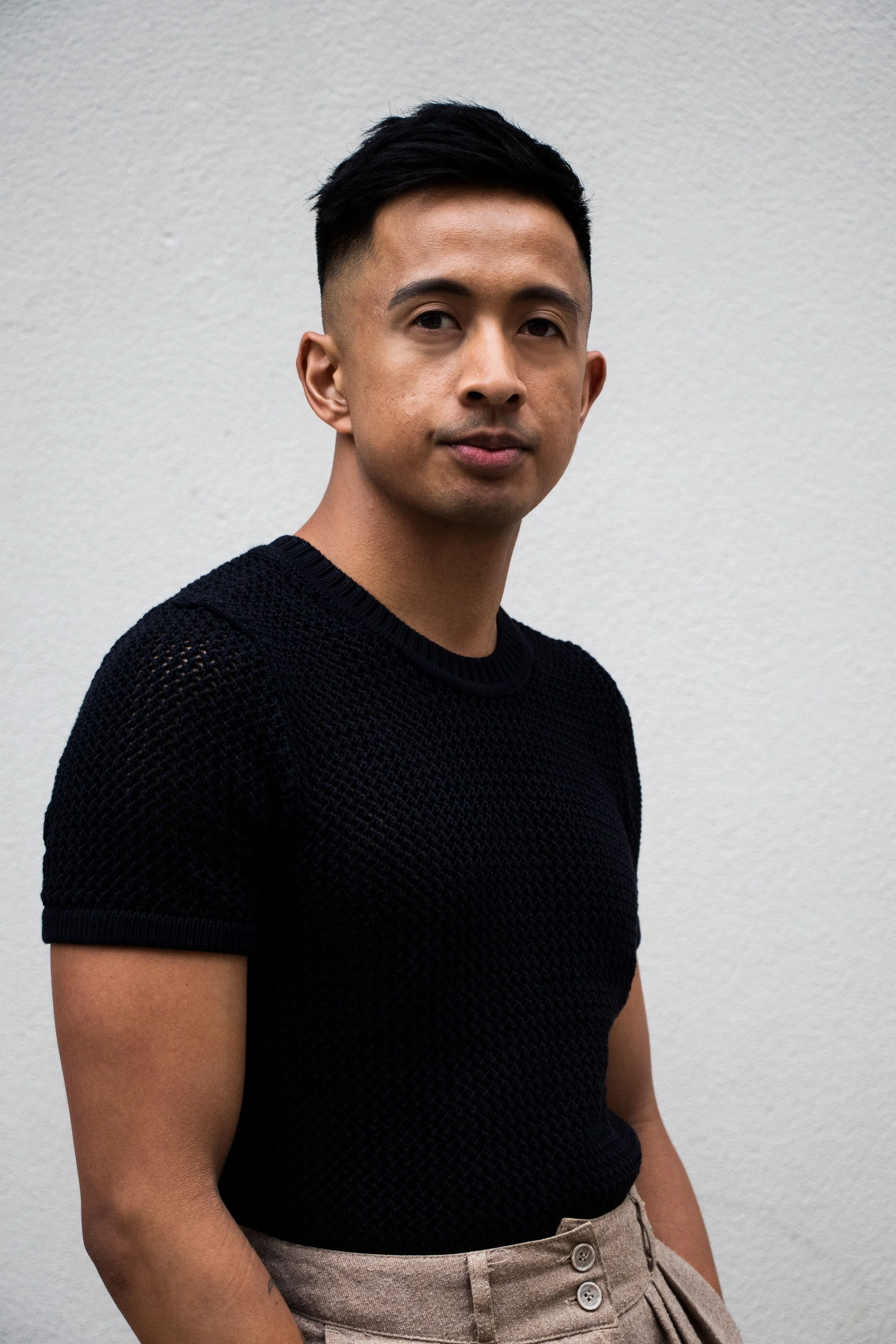 A young man with short black hair, wearing a black textured short-sleeve shirt and beige high-waisted pants, standing against a plain white wall.
