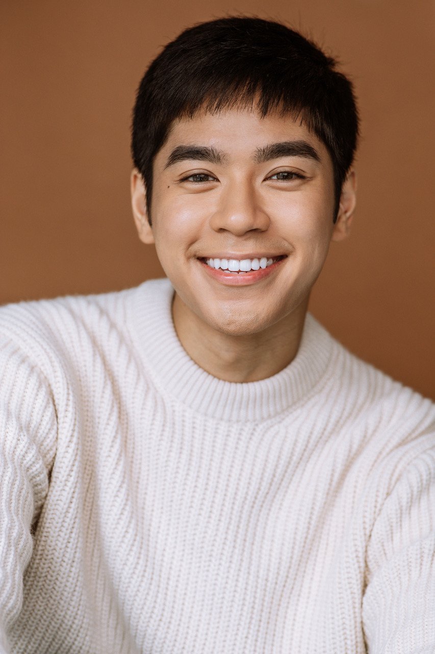 A young man with short black hair, smiling widely, wearing a cream-colored knit sweater, against a brown background.