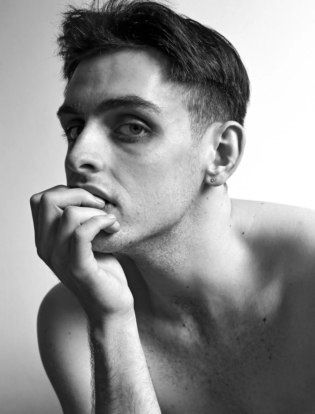 Black and white close-up photo of a young man with short hair, looking directly at the camera with his chin resting on his hand, shirtless, with a piercing in his ear.