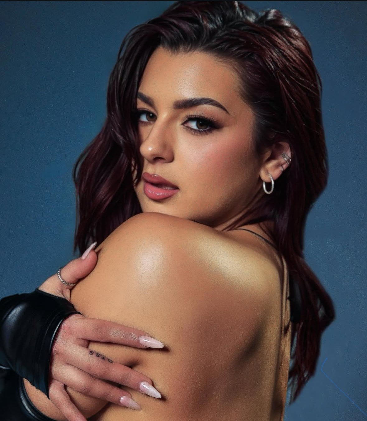 A woman with reddish-brown hair, wearing earrings and a finger ring, with makeup, looking over her shoulder, holding her shoulder with one hand, against a blue background.