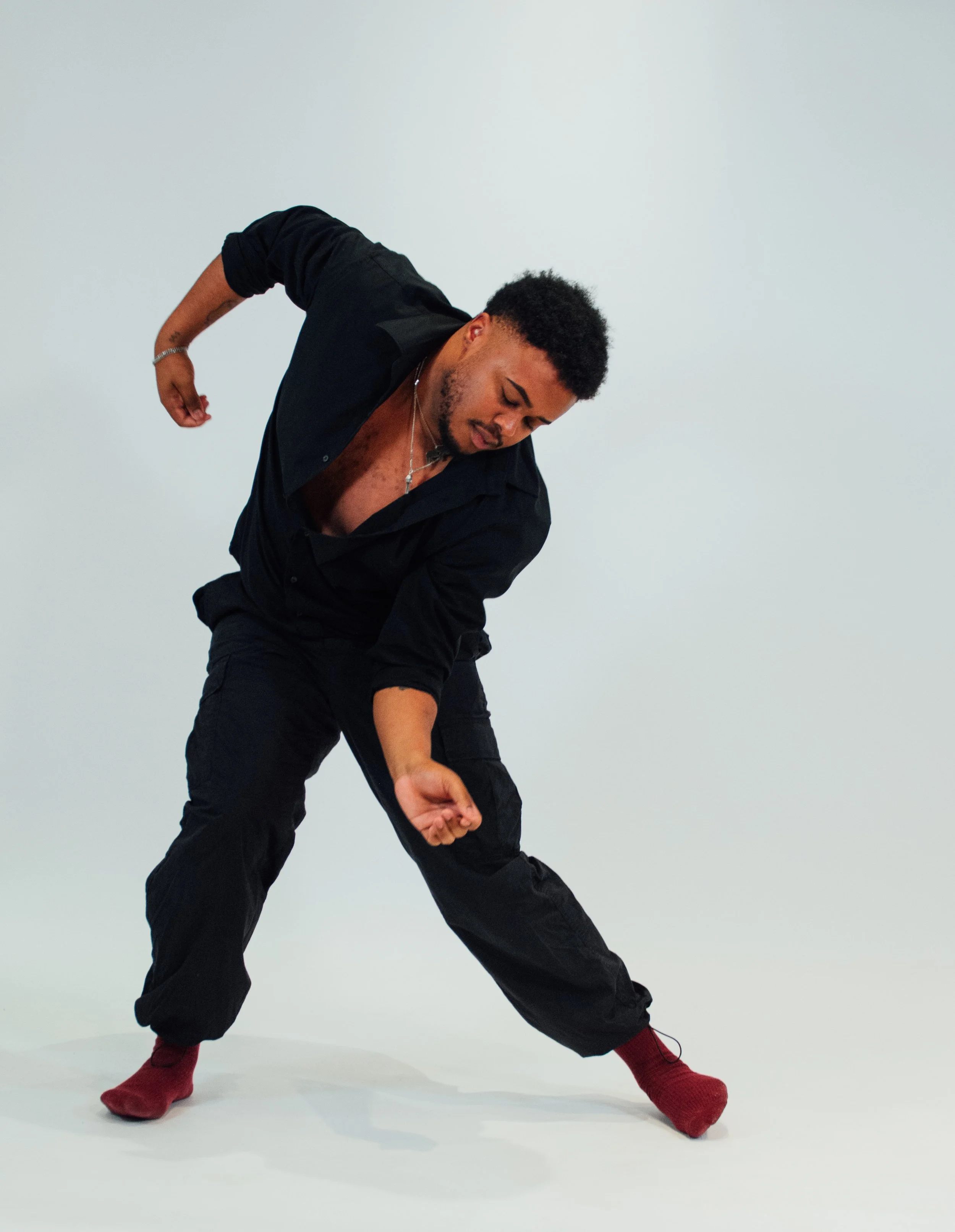 A man in black clothing and red socks bending forward with one arm extended down and the other arm bent behind his back, against a plain white background.