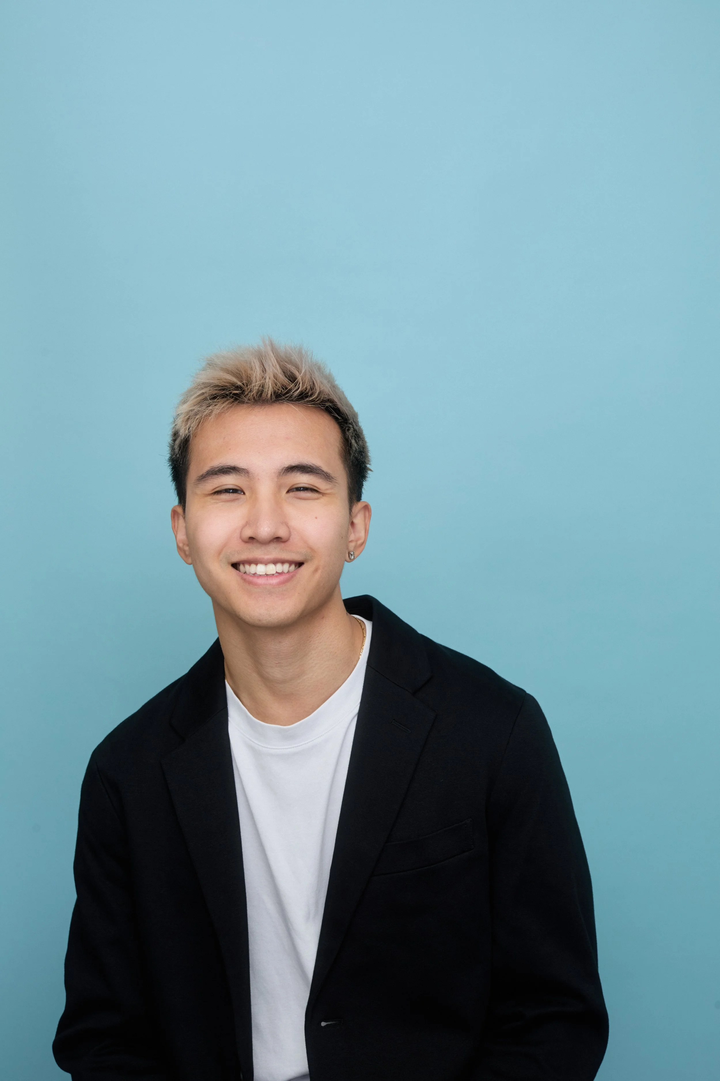 Portrait of a young man with blond-tipped hair smiling, dressed in a black blazer and white t-shirt, against a light blue background.