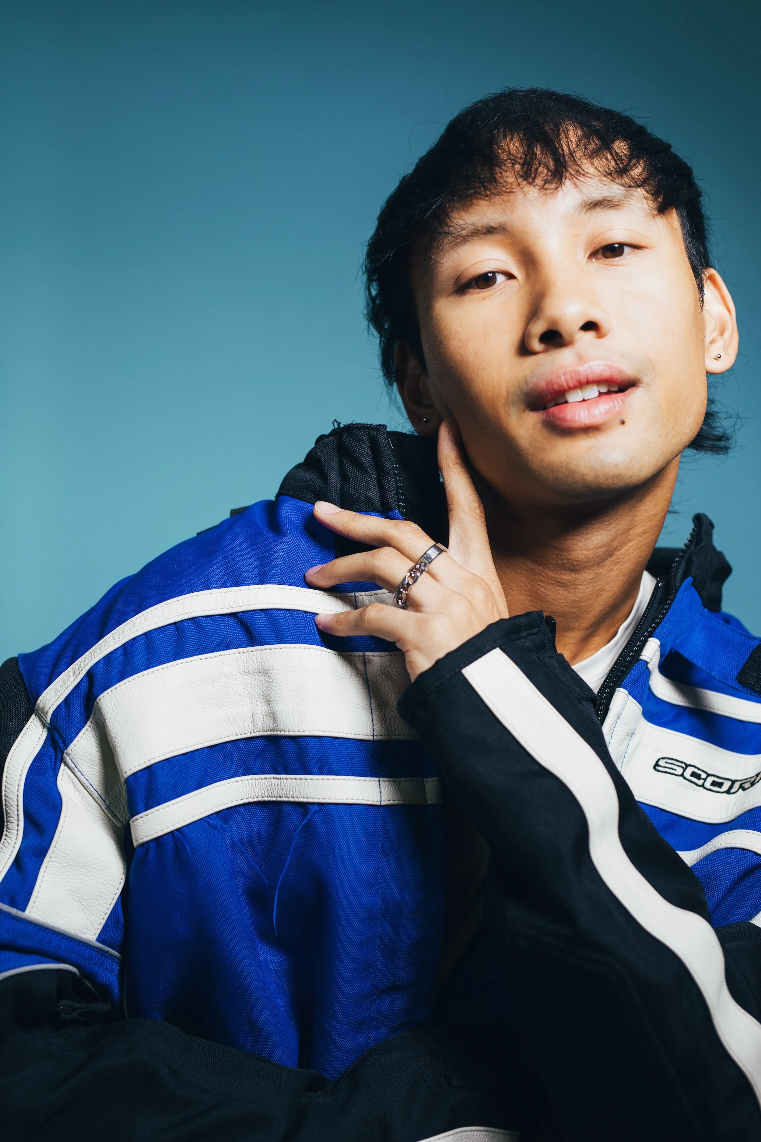 Portrait of a young man with light skin and short dark hair, wearing a blue and white motorcycle jacket, posing with his hand resting on his neck against a plain teal background.