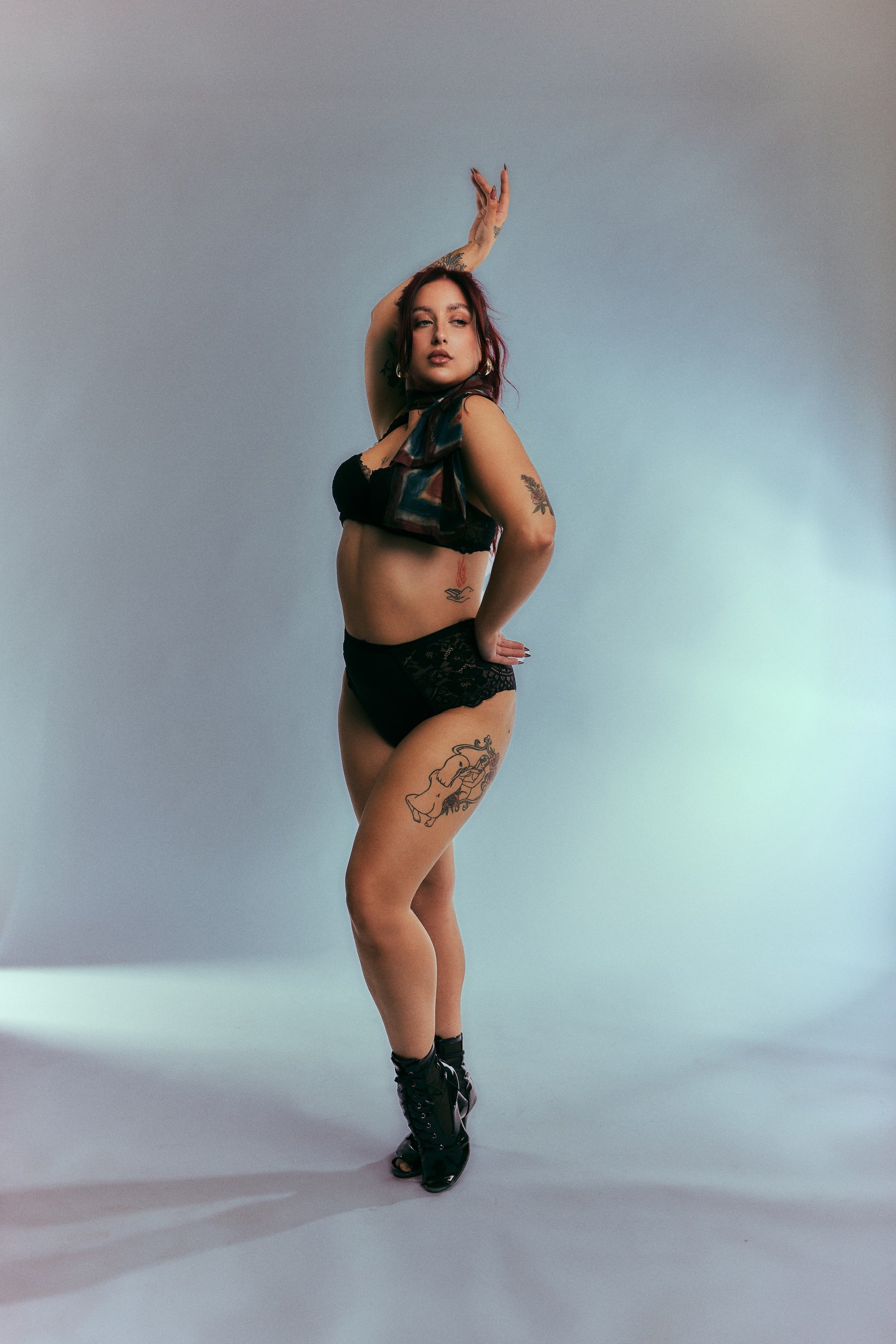 A woman standing in a studio with a plain backdrop, wearing black lace lingerie, high-heeled boots, and posing with one arm raised above her head and the other on her hip. She has colorful tattoos on her arms and legs.