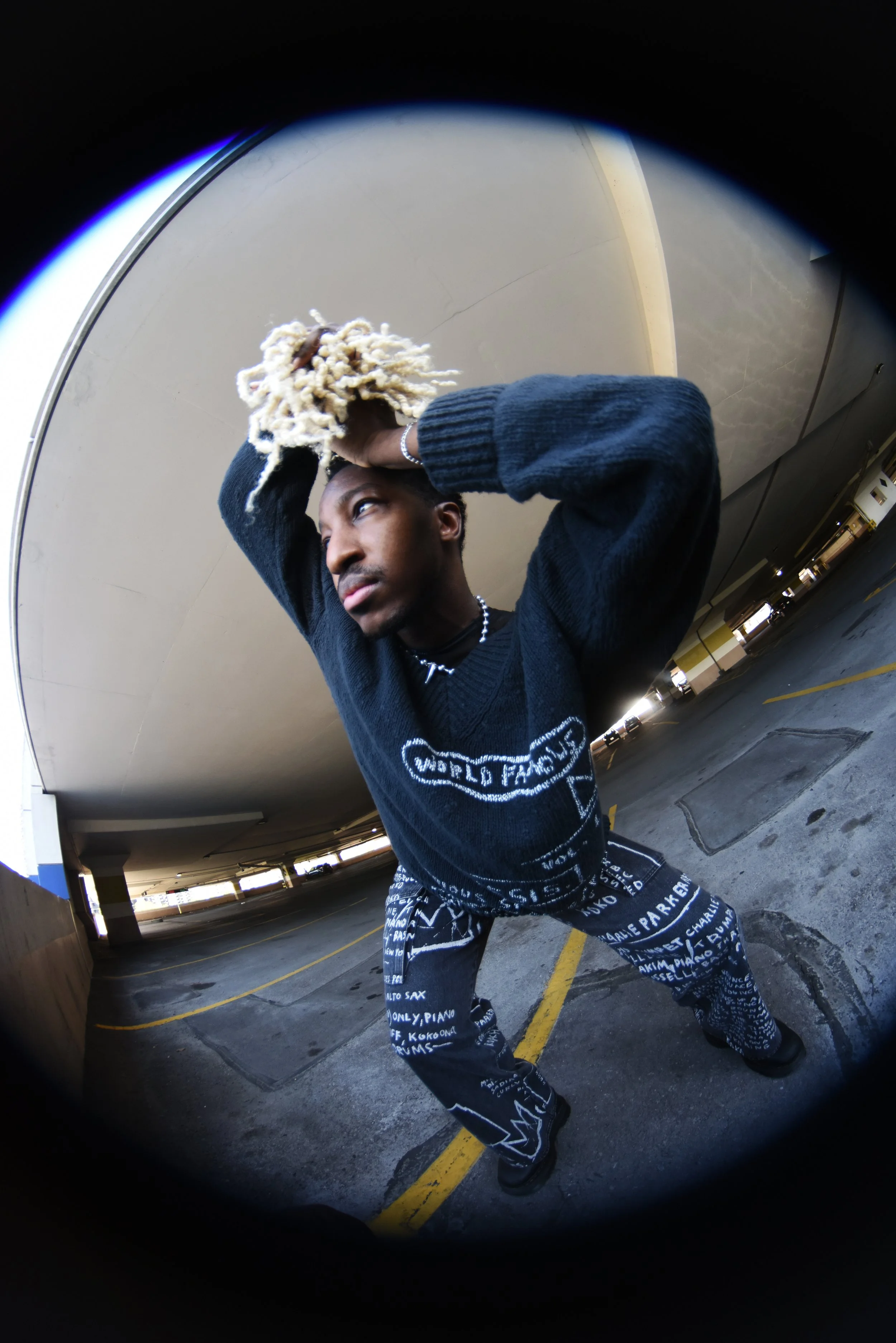 A young man with dreadlocks poses with his hands on his head in a parking garage, wearing a black sweater and patterned pants with writing on them, taken with a fisheye lens.