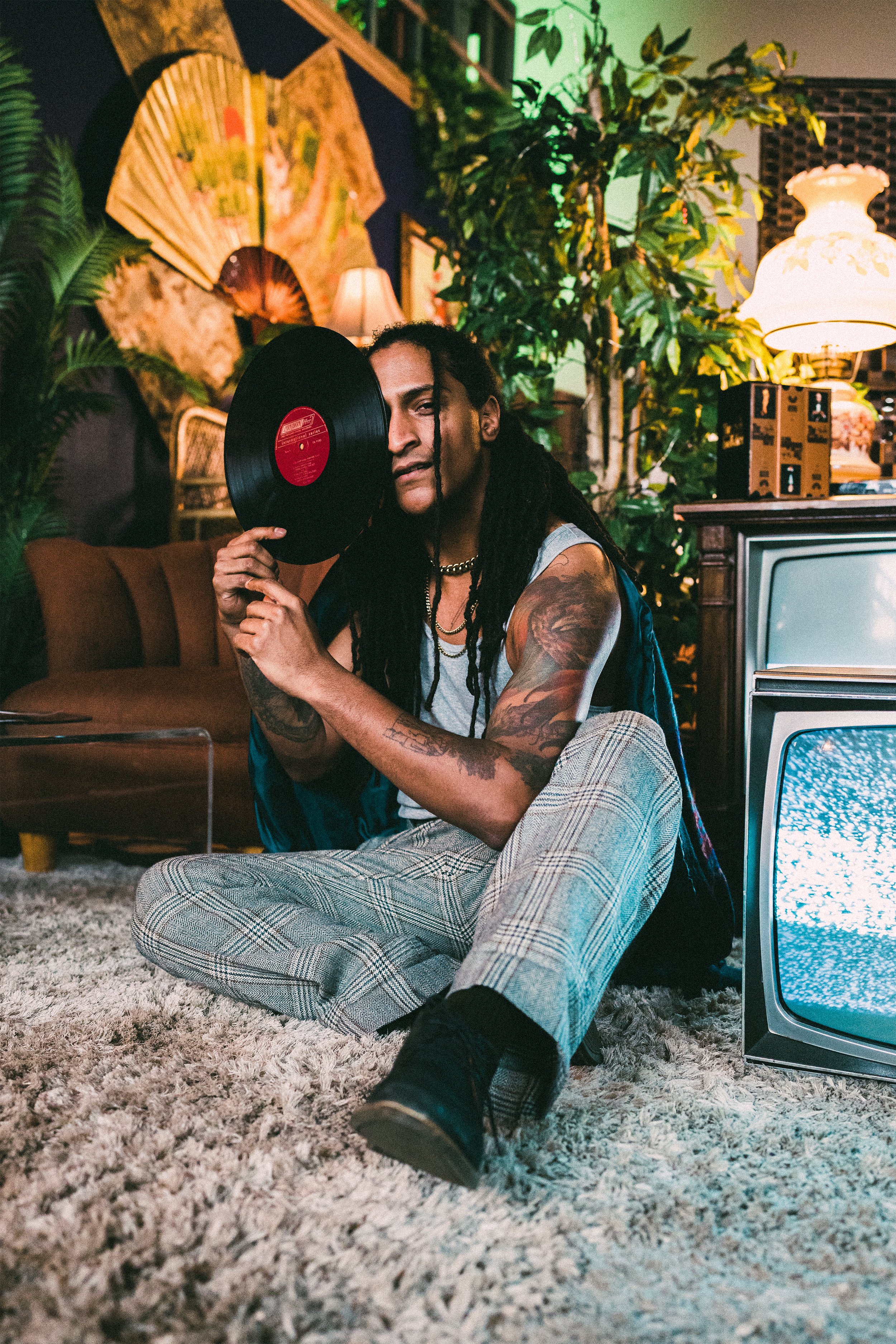 A person with long dreadlocked hair and tattoos on their arms is sitting on a shaggy rug in a vintage-themed room, holding a vinyl record up to their face. The room features lush green plants, a standing lamp, decorative fans on the wall, and an old television with static on the screen.