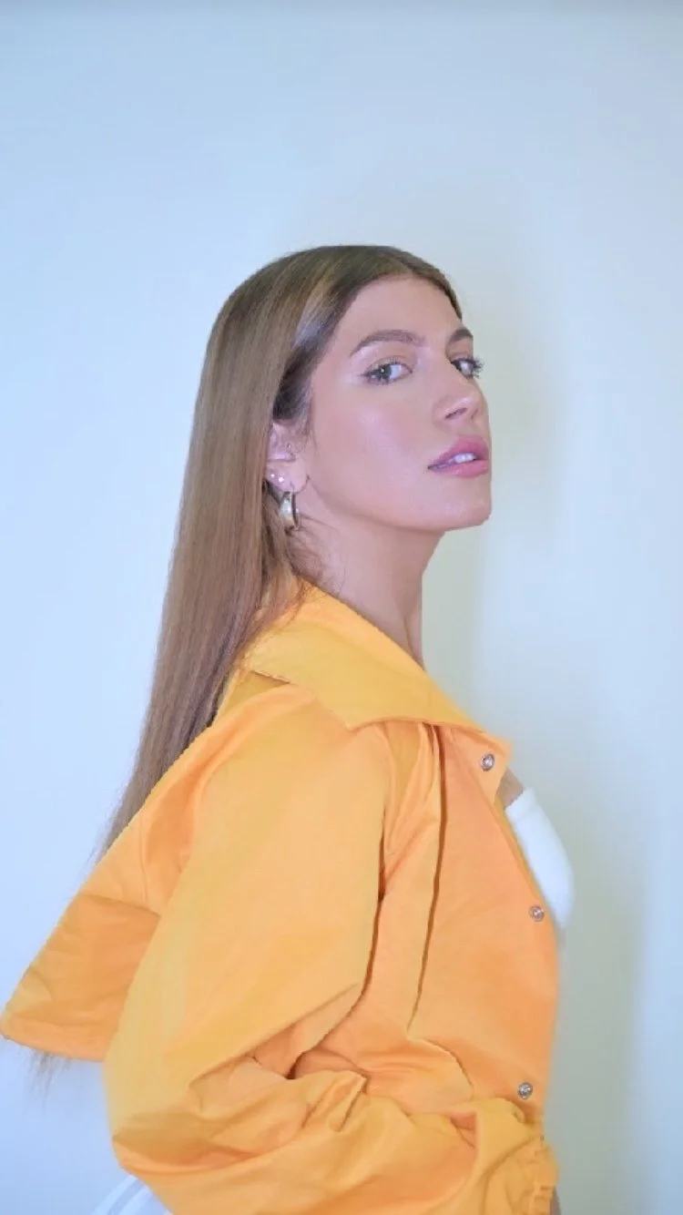 A woman with long, straight, light brown hair wearing a yellow jacket, looking over her shoulder at the camera, with a neutral background.