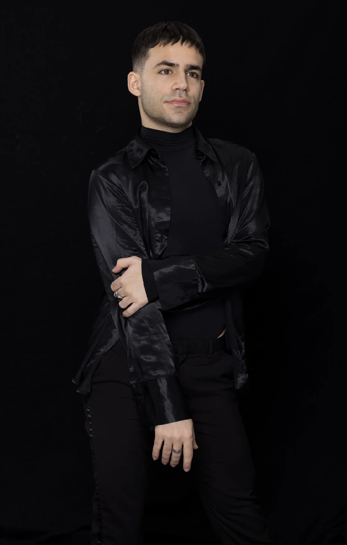 Portrait of a young man with short dark hair, wearing a black turtleneck, black shiny jacket, and black pants, posing against a black background.