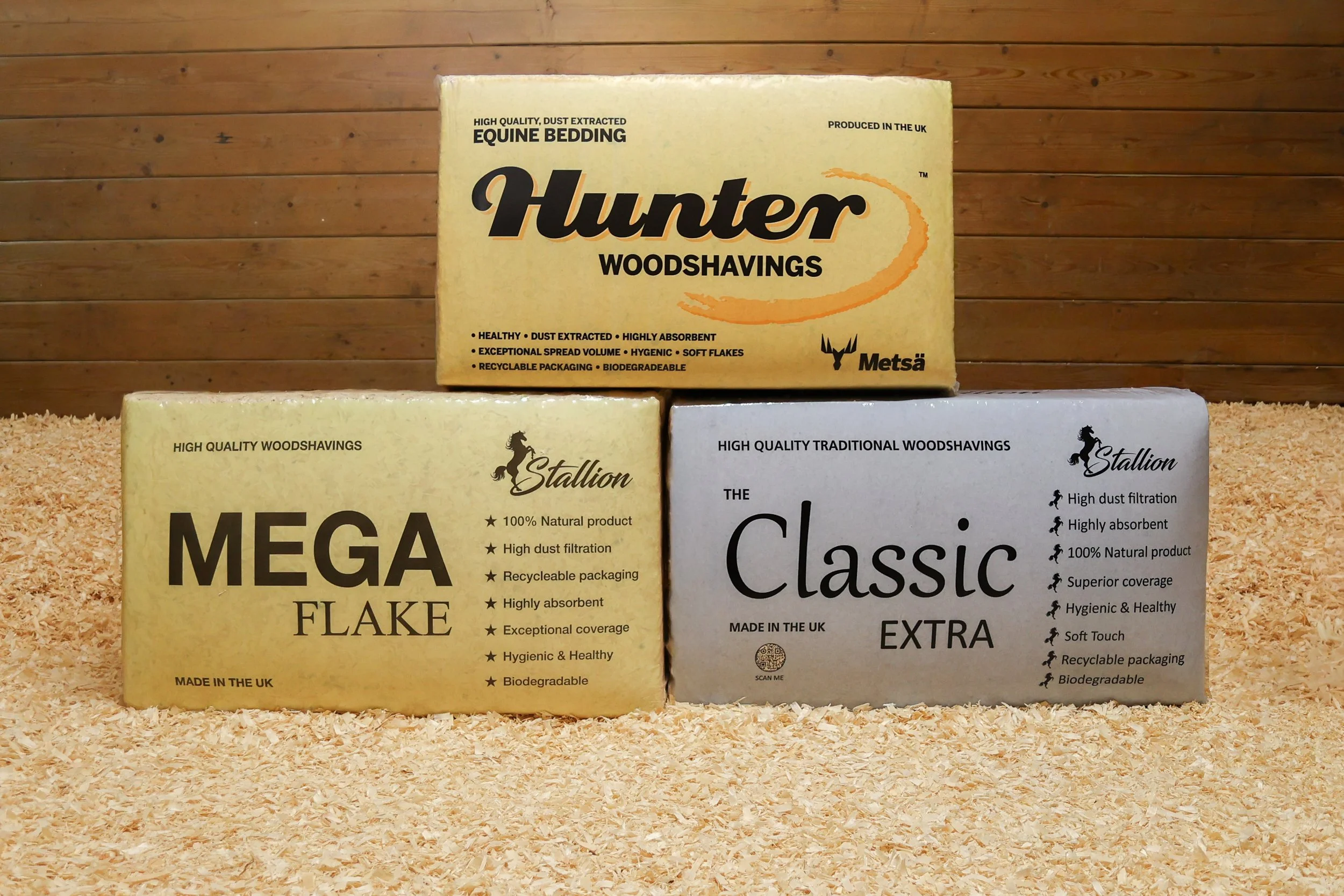 Boxes of woodshavings for equine bedding and traditional woodshavings, placed on a bed of wood shavings against a wood-paneled wall.