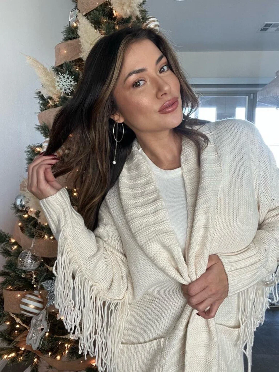 Woman wearing a white fringed sweater poses in front of a decorated Christmas tree.