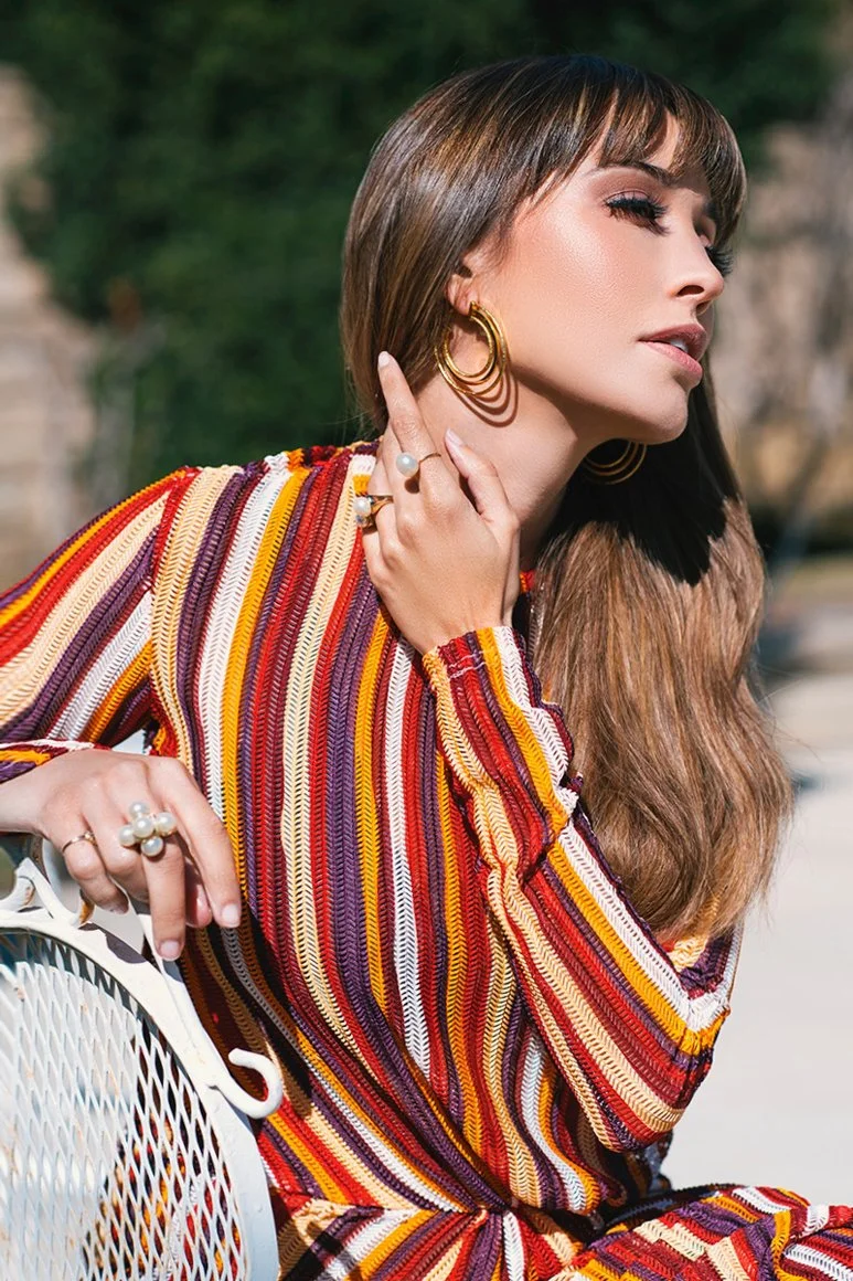 Woman with long hair, wearing a colorful striped outfit and gold hoop earrings, sitting on a white metal chair outdoors.