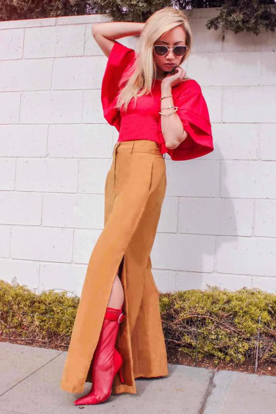 Fashionable woman wearing red top, tan pants, and red high-heeled boots, posing against a white brick wall.