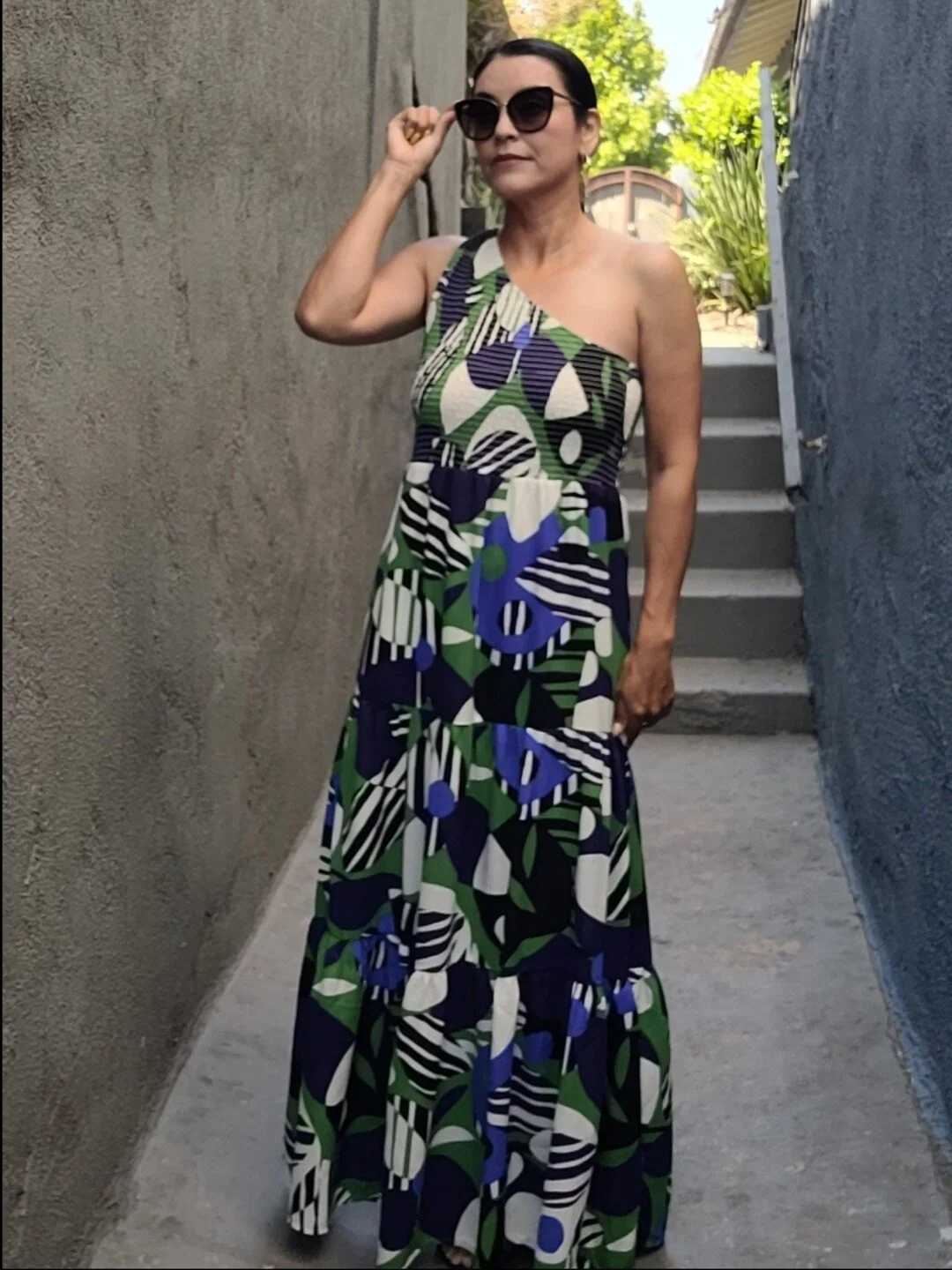 Woman wearing a geometric-patterned, one-shoulder dress and sunglasses in a narrow alleyway.