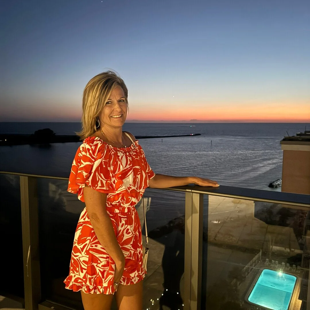 Woman in red and white dress standing on a balcony with ocean view and sunset in the background.
