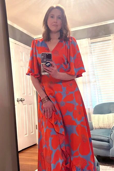 Woman taking a mirror selfie wearing a long, floral dress with red and blue colors in a room with a door, window, and chair in the background.