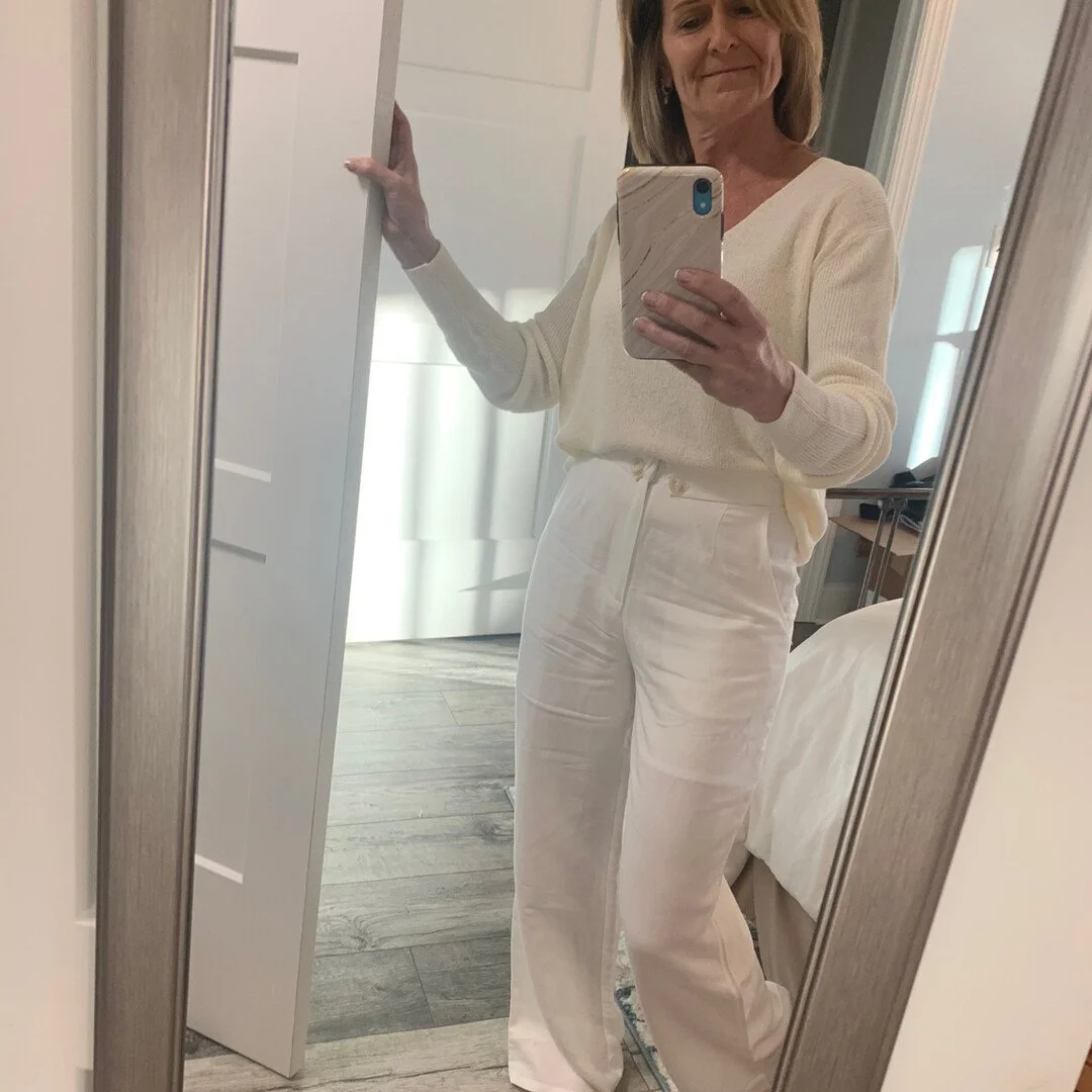 Person taking a mirror selfie wearing a white sweater and white pants in a bedroom.
