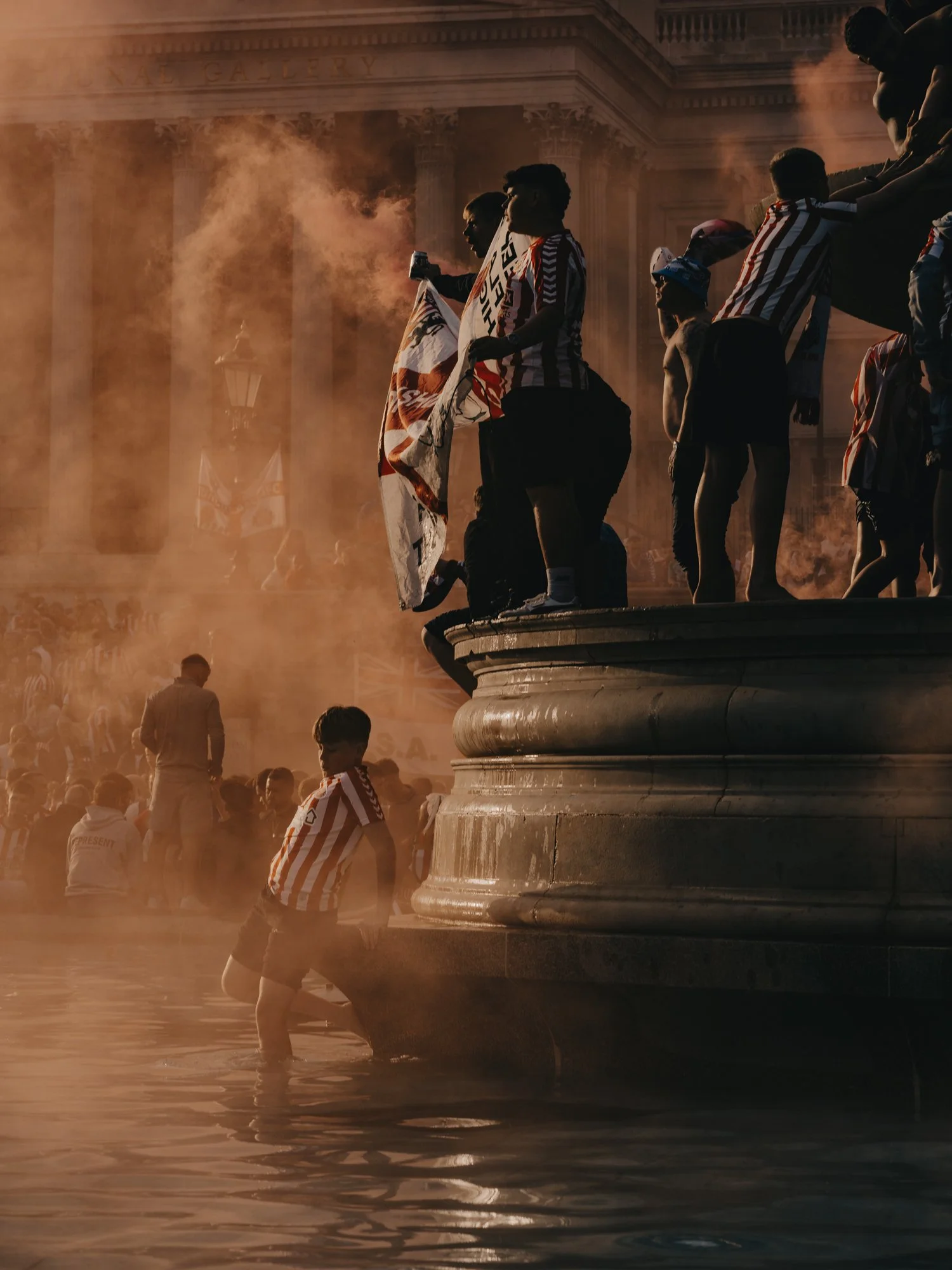 Moody, cinematic street photography in smoke filled Trafalgar Square London. Football fans seen through dense smoke on the fountain by Joshua K Jackson.