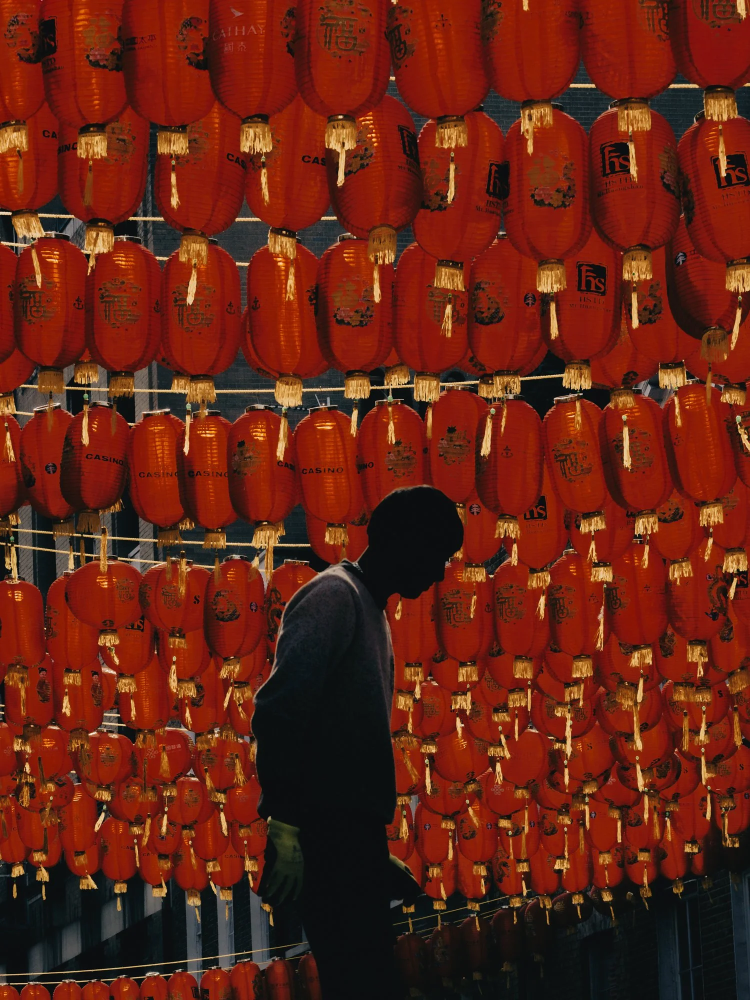 A lone human silhouette is positioned in front of a dense wall of hundreds of illuminated red paper lanterns. High contrast and deep colors in London Chinatown by Joshua K Jackson.