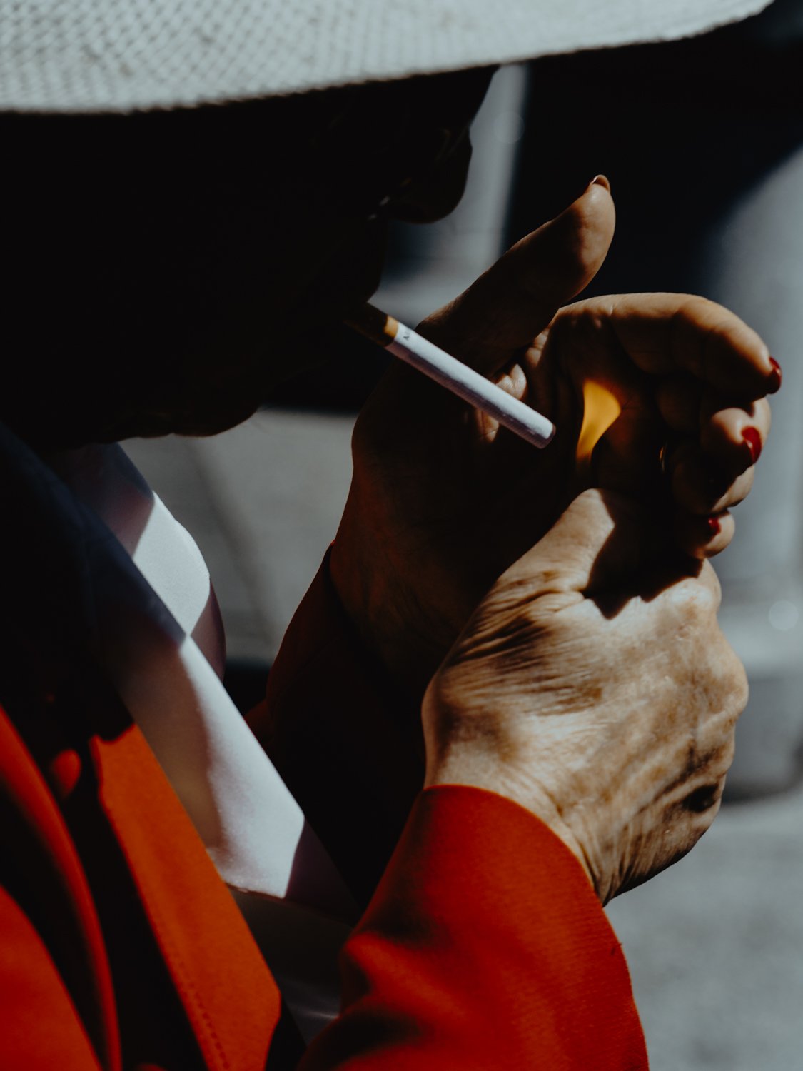 Close-up of a person in a red coat and wide-brimmed hat lighting a cigarette, London street photography by Joshua K. Jackson.