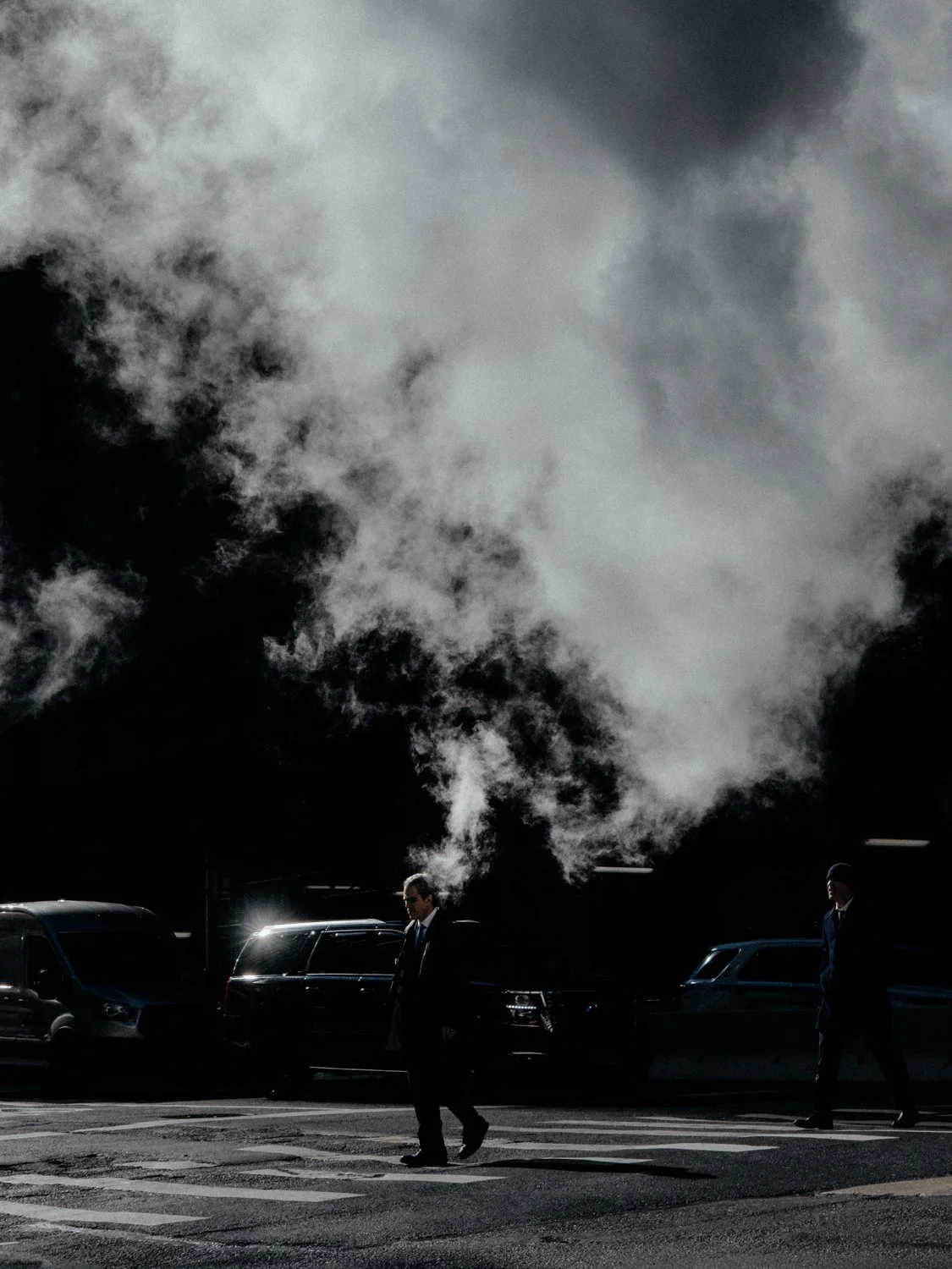 Man crossing a New York street beside rising steam and parked cars at night, street photography by Joshua K. Jackson.