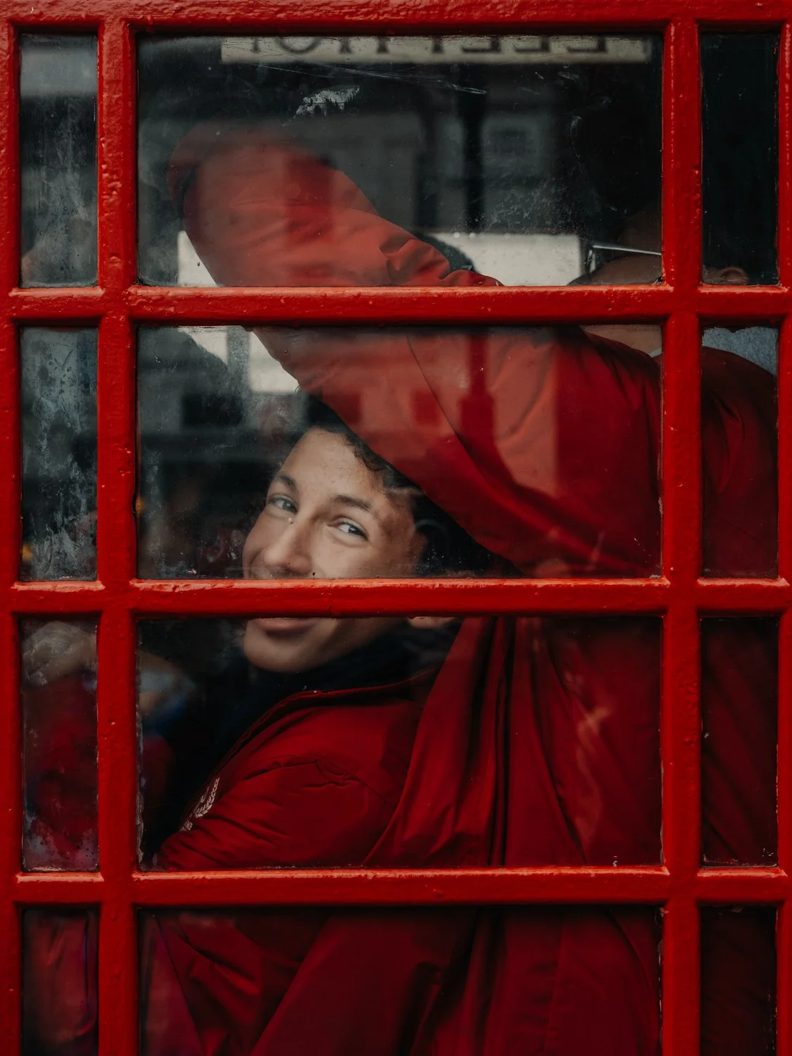 Man in a red coat smiling through a red Covent Garden London telephone box, street photography by Joshua K. Jackson.