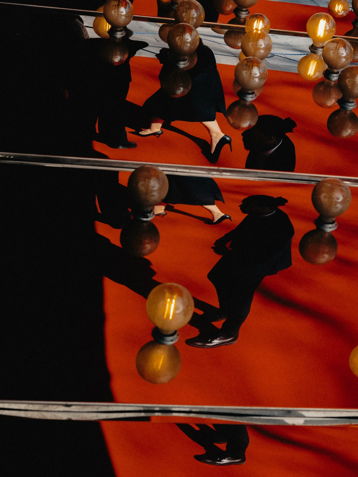 Graphic, abstract street photography. Strong directional light casts dramatic human silhouettes and geometric shadows from golden spheres onto a bright red walkway by Joshua K Jackson.