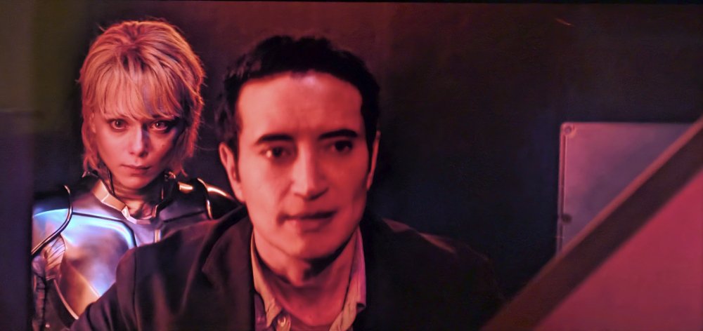 Screenshot from M3GAN 2.0 showing Christian standing in an elevator, intending to escape. Amelia stands behind him, a viper poised to strike.