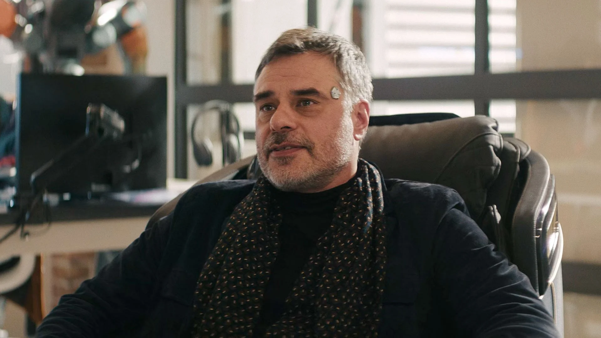 Screenshot of the upper body of tech billionaire Alton Appleton from M3GAN 2.0. He's sitting in his futuristic wheelchair and a visible neurochip is attached to his left temple.