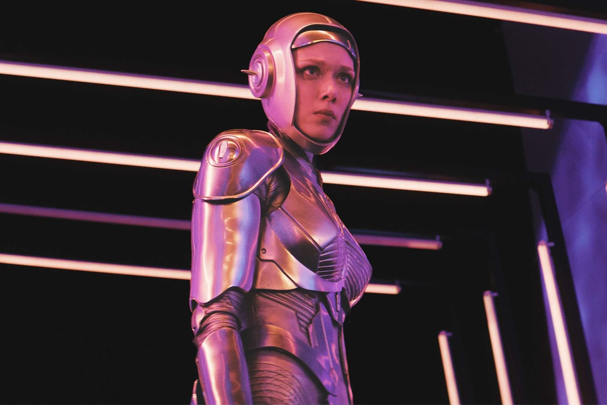 Screenshot of Amelia from M3GAN 2.0.  She's wearing a metallic, form-fitting outfit and standing against rows of neon lights. She wears a helmet and her white face is visible.
