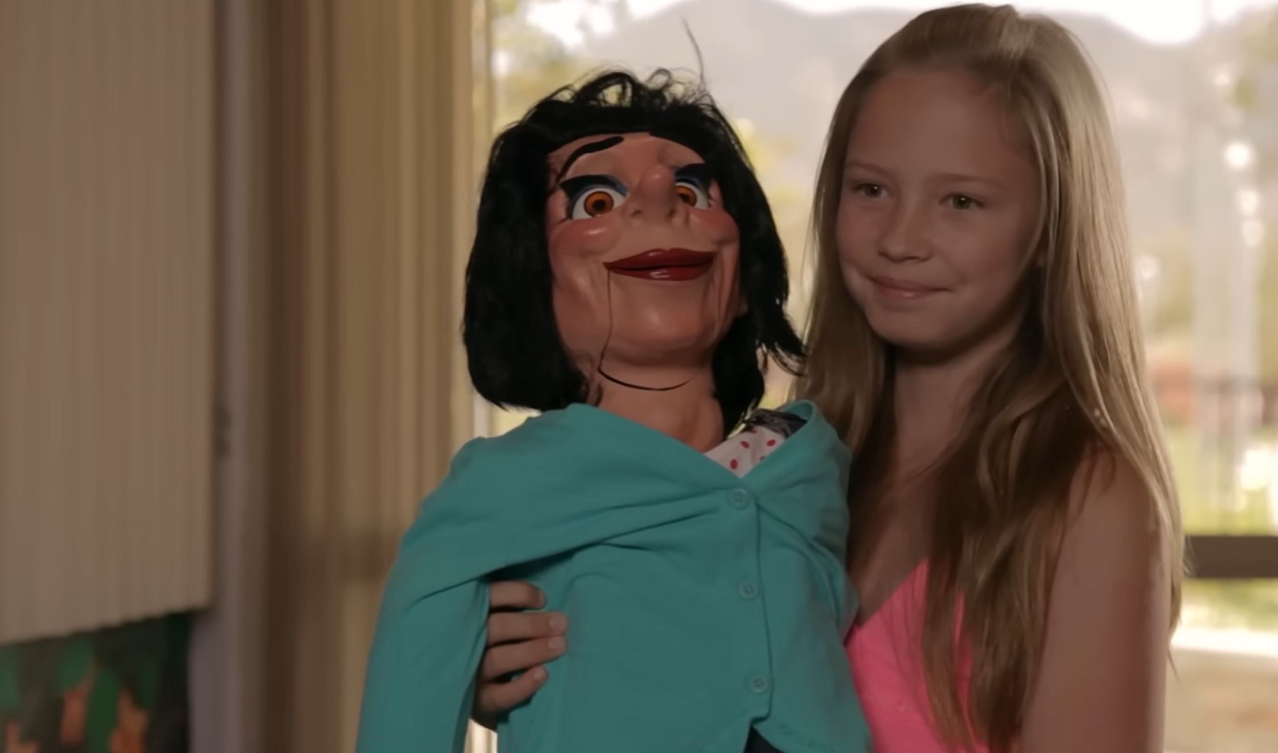Screenshot of the doll Miss Rachel and Lacy, a white, eight year old girl from the 2012 haunted doll film Paranormal Adoption. Lacy holds her doll while meeting her new adoptive parents.