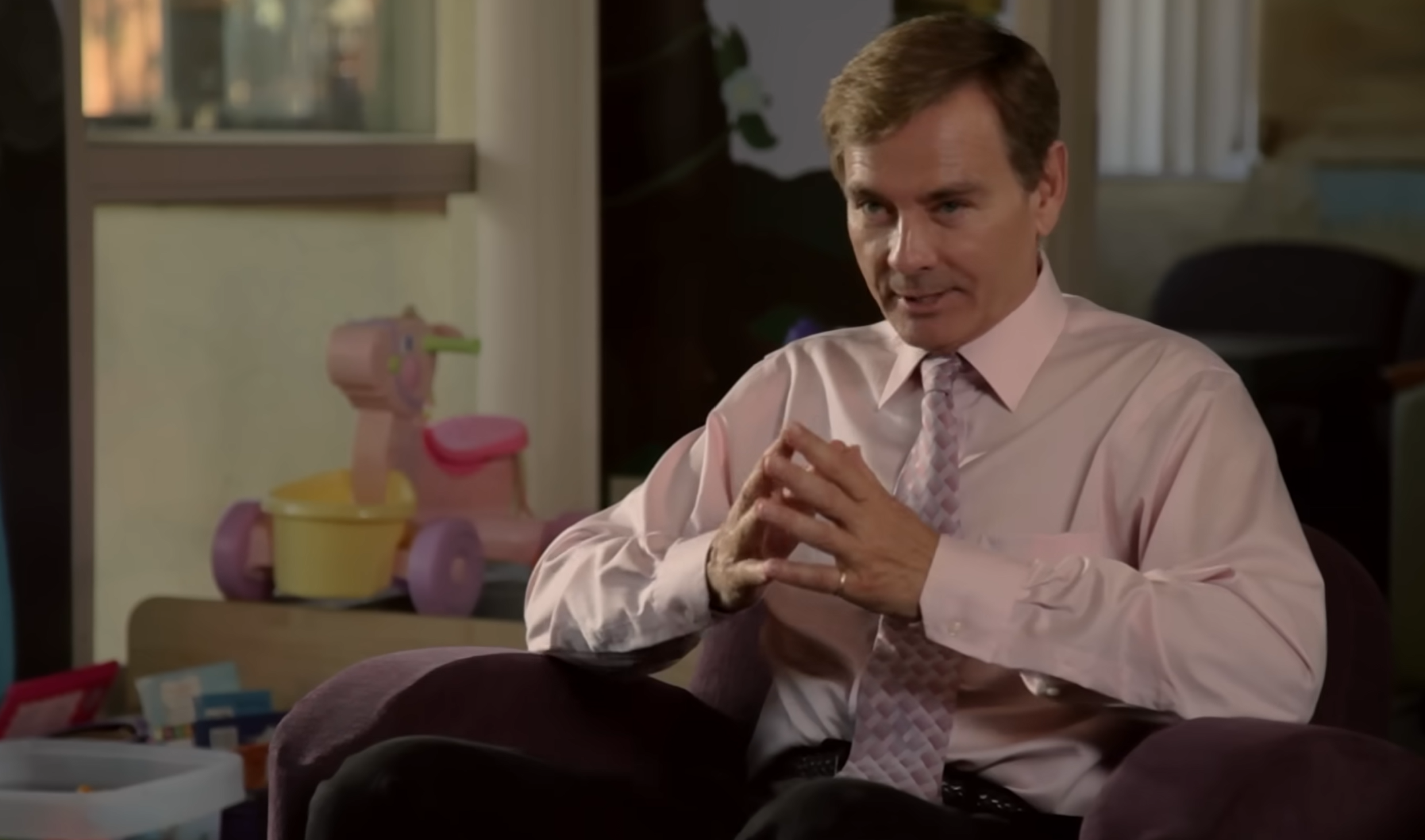 Screenshot of the adoption agency owner from the 2012 haunted doll film Paranormal Adoption. He's sitting in an office while addressing a prospective adoptive couple. He's a white man wearing a dress shirt and tie. He's steepling his hands.