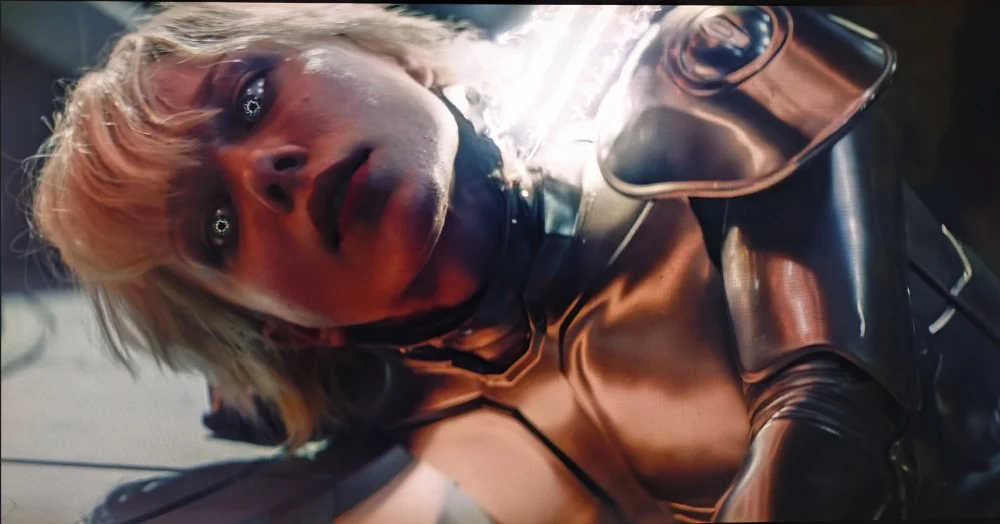 Screenshot of the android Amelia from M3GAN 2.0. She's on the ground on her side as her attackers shock her with a futuristic taser. Her eyes glow silver, indicating her system is being harmed.