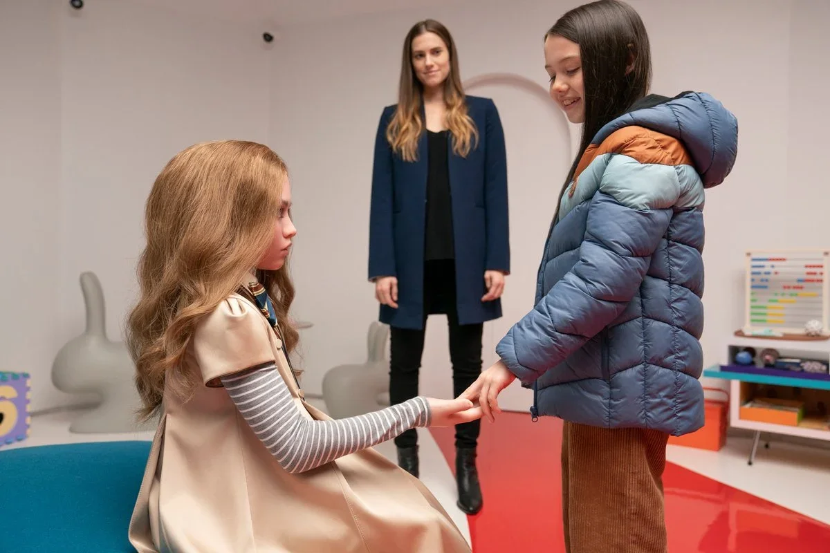 Screenshot from the sci-fi horror film M3GAN. Cady is bonding with M3GAN by pressing her fingers into M3GAN's palm as Gemma looks on from the background.