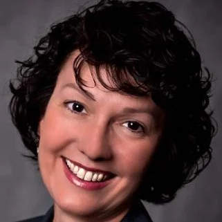 Headshot of Lynette Ward, Coach, smiling with short, curly dark hair, smiling with teeth showing, against a dark gray background.