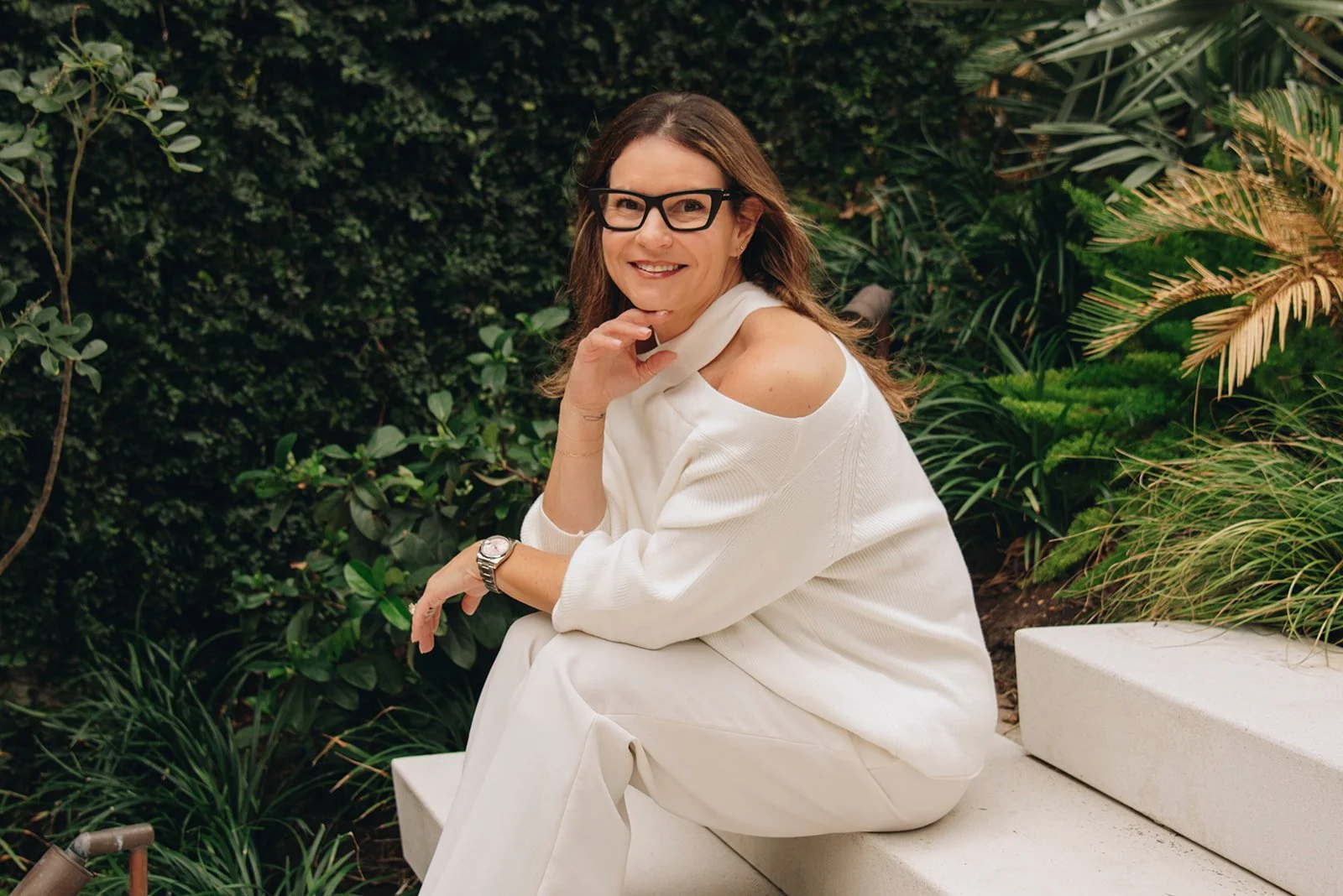 A woman with glasses and brown hair sitting on a white bench outdoors, smiling at the camera, surrounded by green plants.