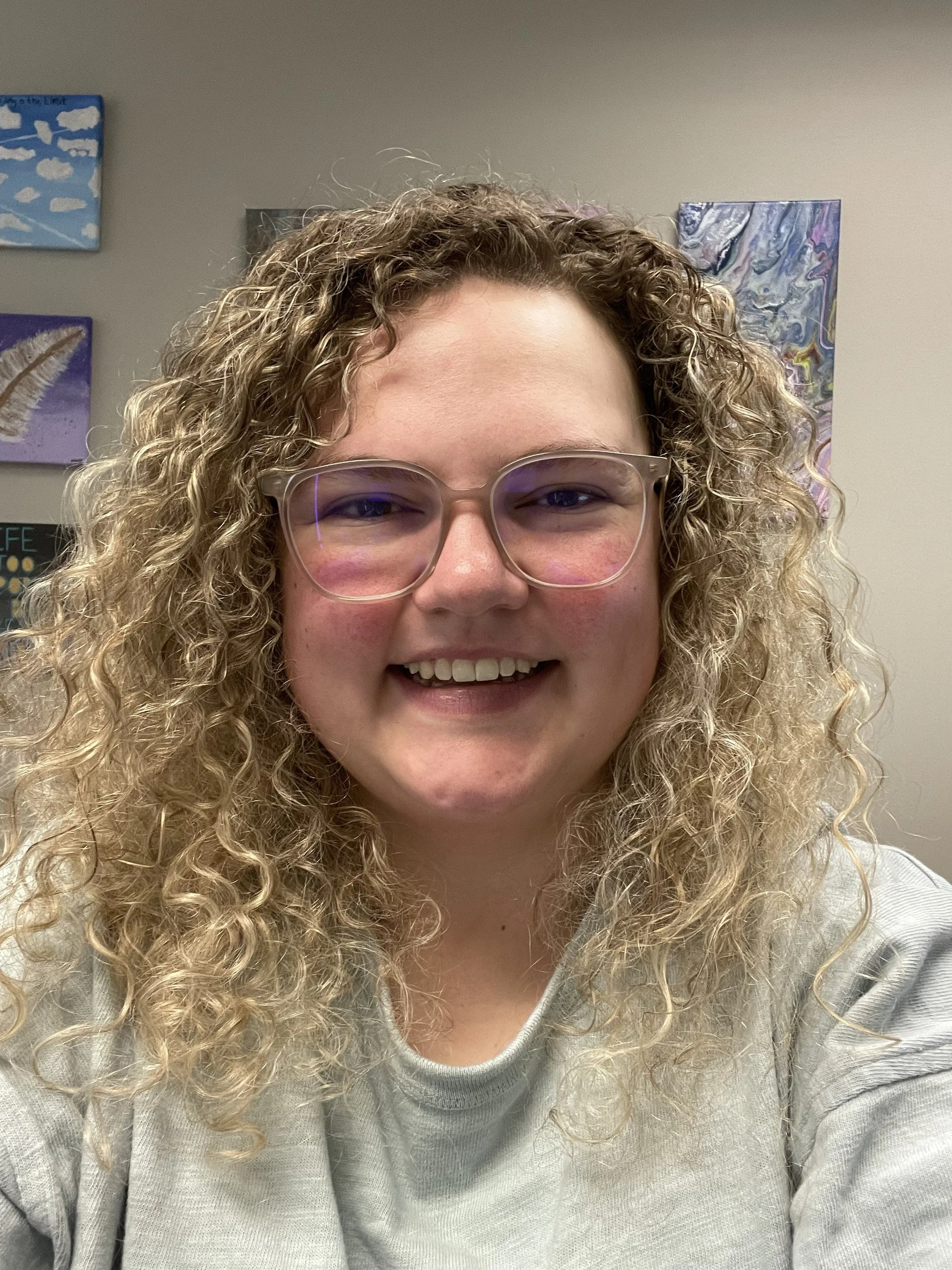 A young woman with curly blonde hair wearing glasses, smiling at the camera in an indoor setting with colorful wall art in the background.