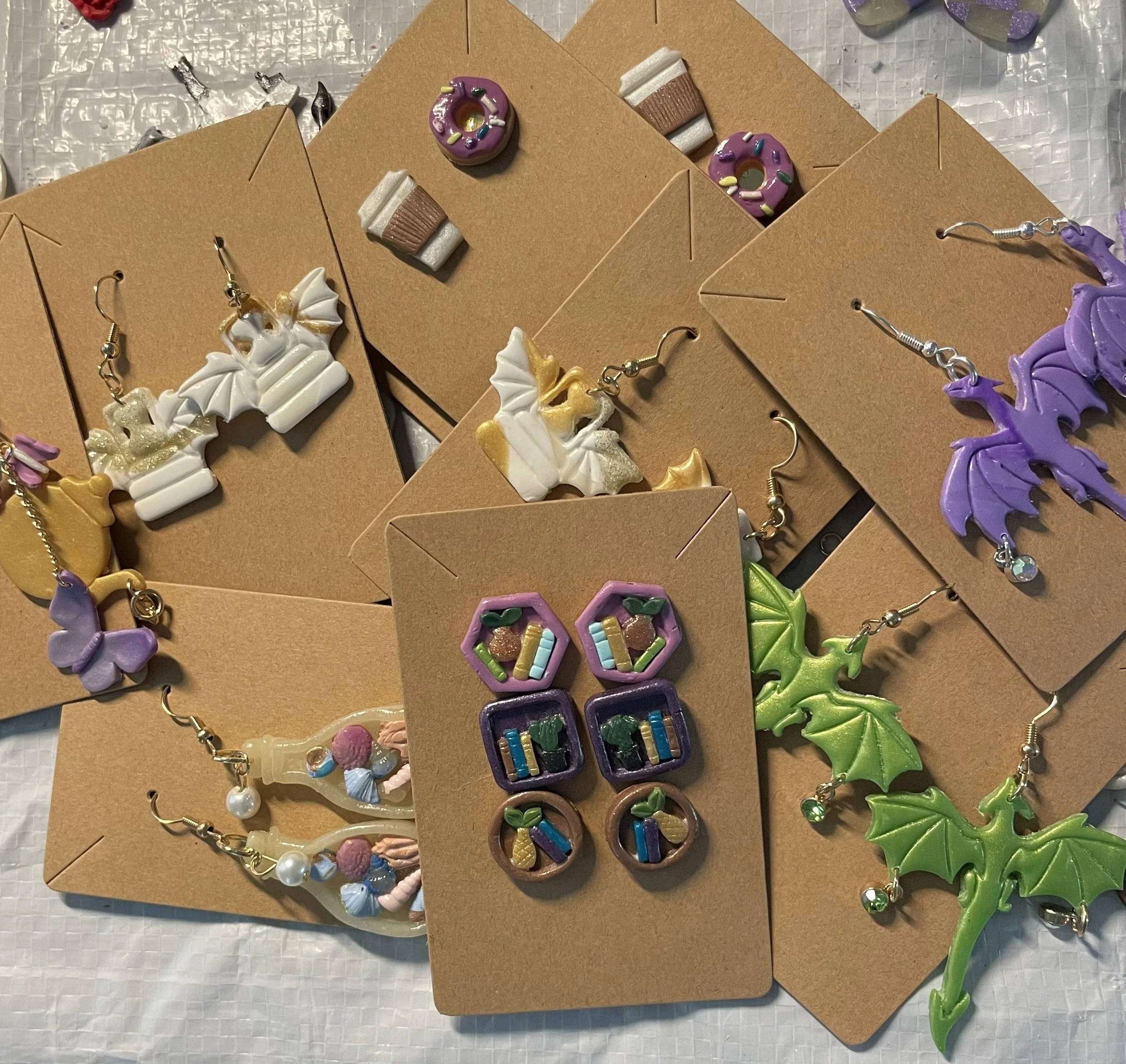 Colorful earrings displayed on brown cardboard backing, including designs like dragonwings, books, dragons, and donuts, arranged on a textured surface.