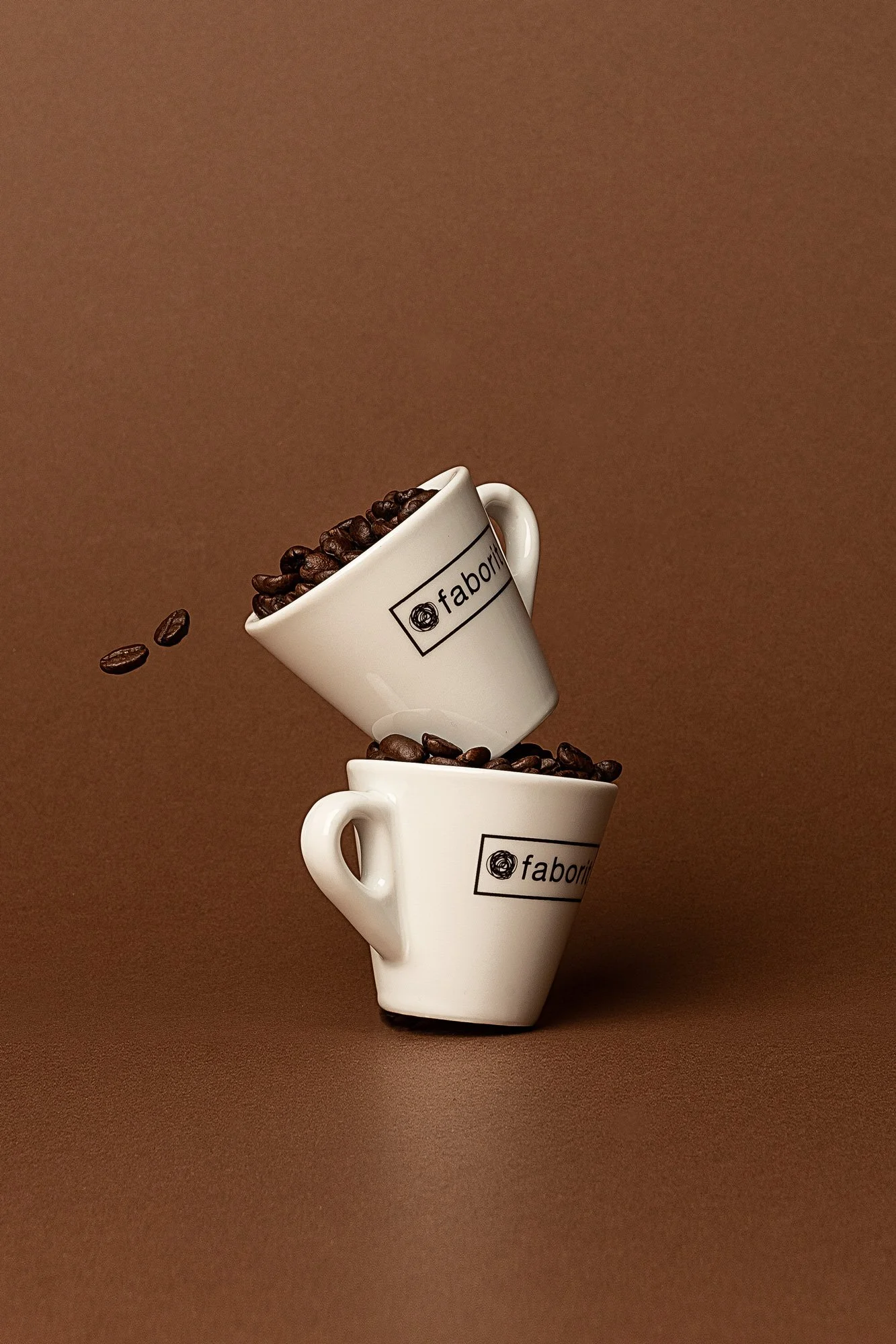 Dos tazas blancas con el logo 'fabori' apiladas, una llena de granos de café y la otra vacía, con algunos granos cayendo al aire.
