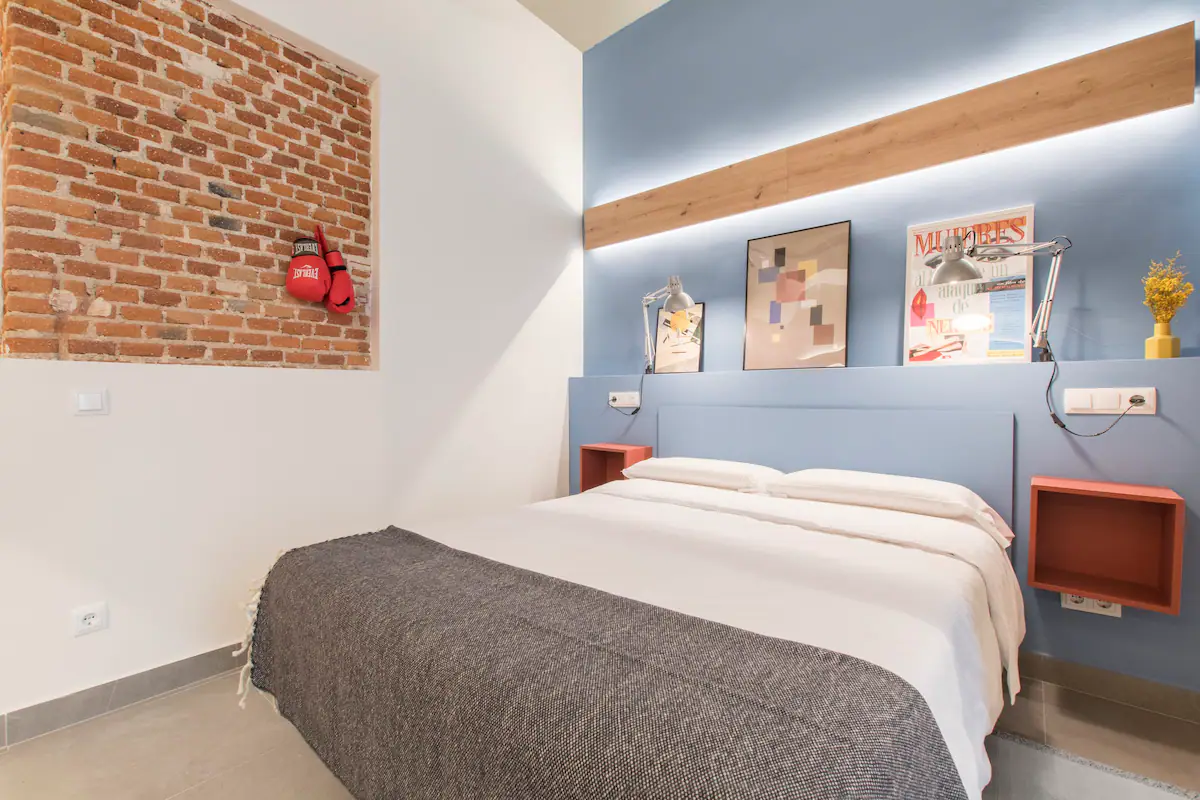 Habitación con cama, paredes blancas y azules, rincón de ladrillos expuestos con guantes de boxeo colgados, arte decorativo y lámparas de lectura